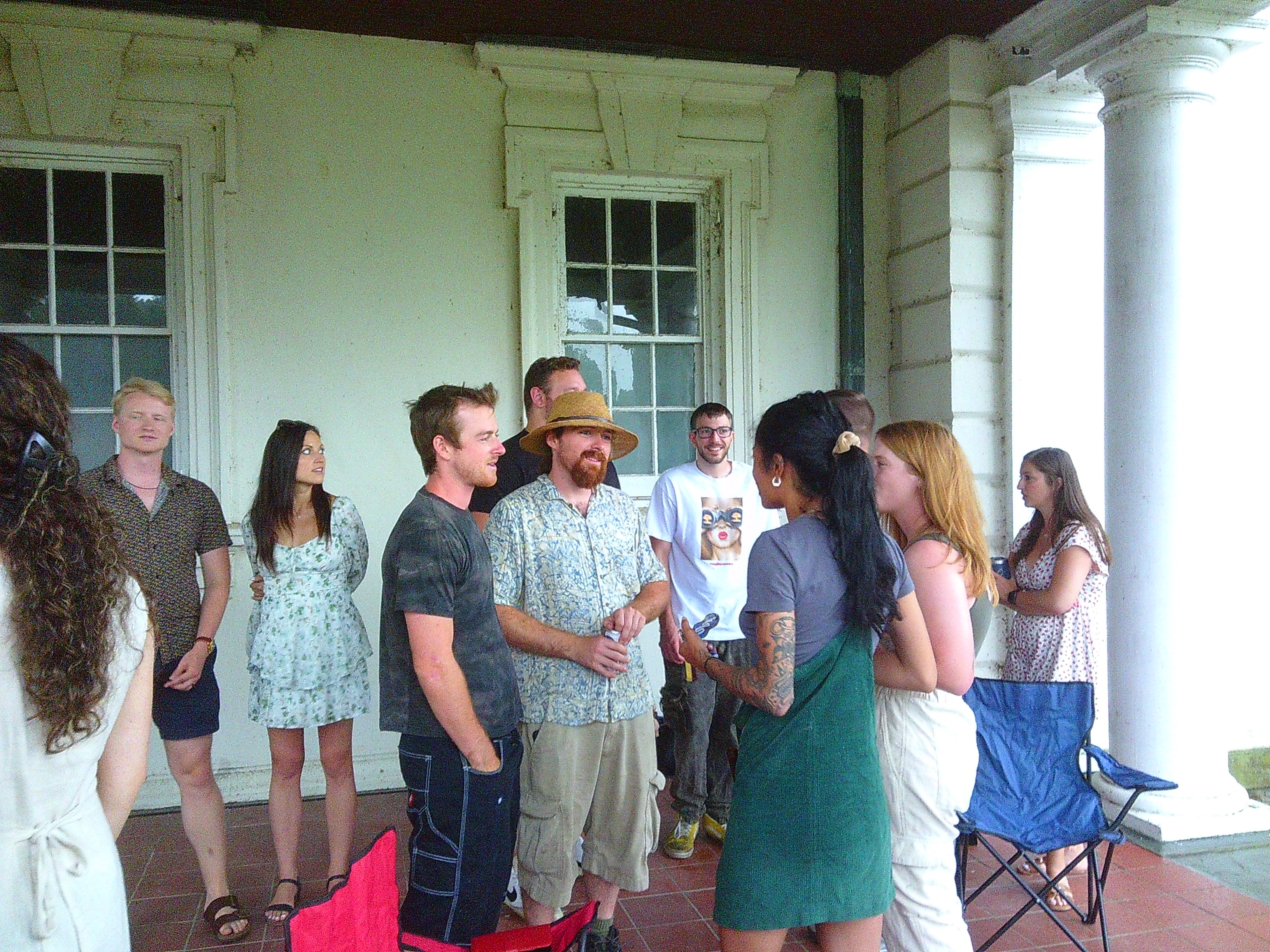 A phot of a group of people standing outside but underneath an overhang with a tiled floor. In the center of the photo, there are 4 people talking to each other, one in a hawaiian shirt as well as a straw hat