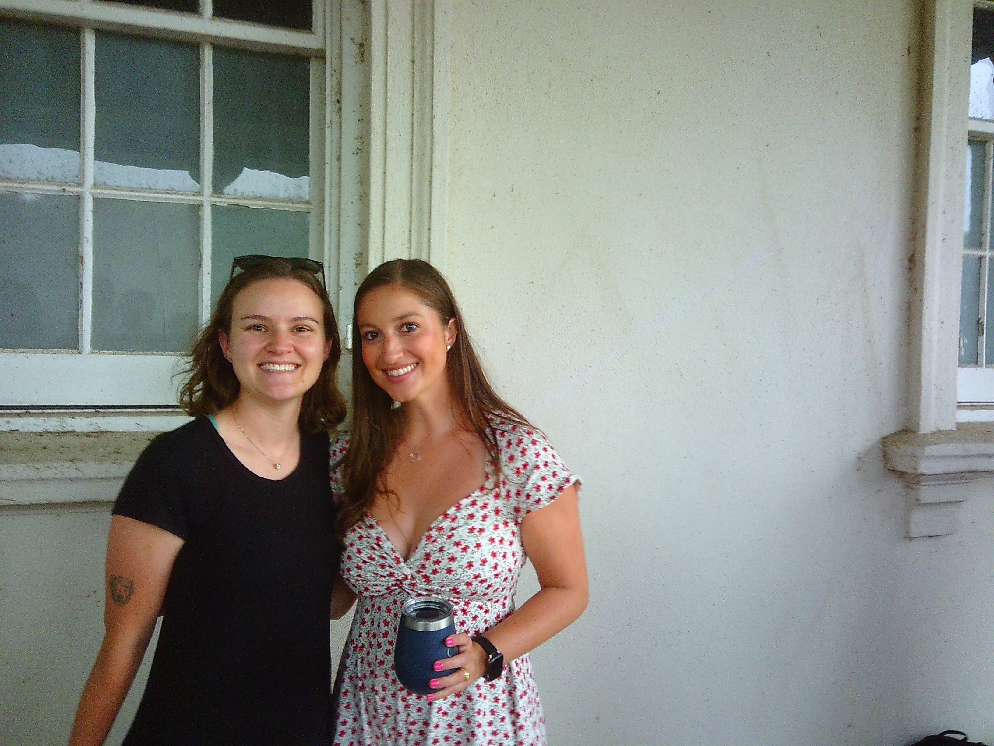 Photo of two women smiling towards the camera. One is in a black dress, the other a sort of white dress with red looking star shaped objects