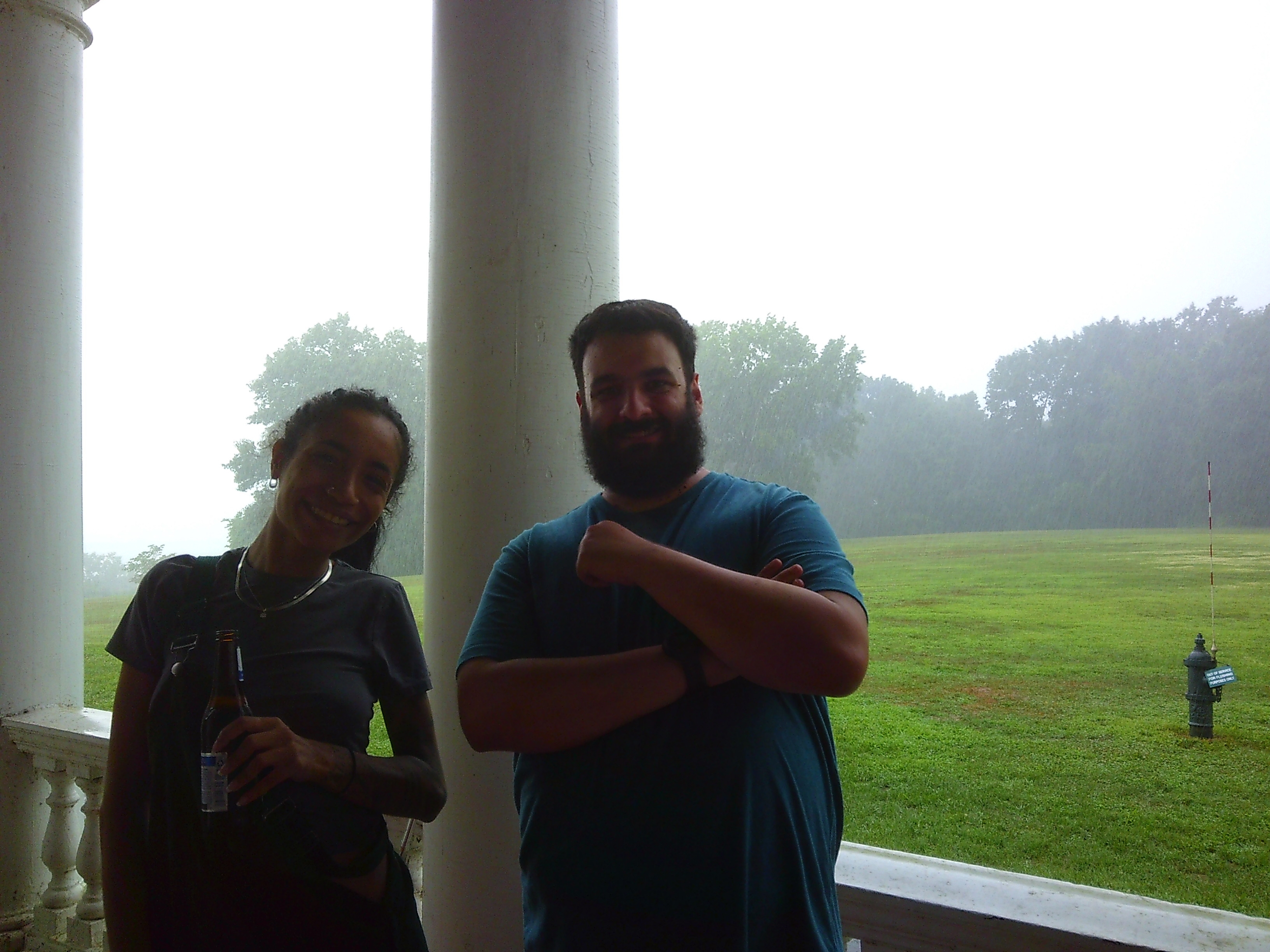 Photo of a man and woman smiling towards the camera. The man is in a blue shirt, the woman in a grey shirt and green overalls. It is raining behind them