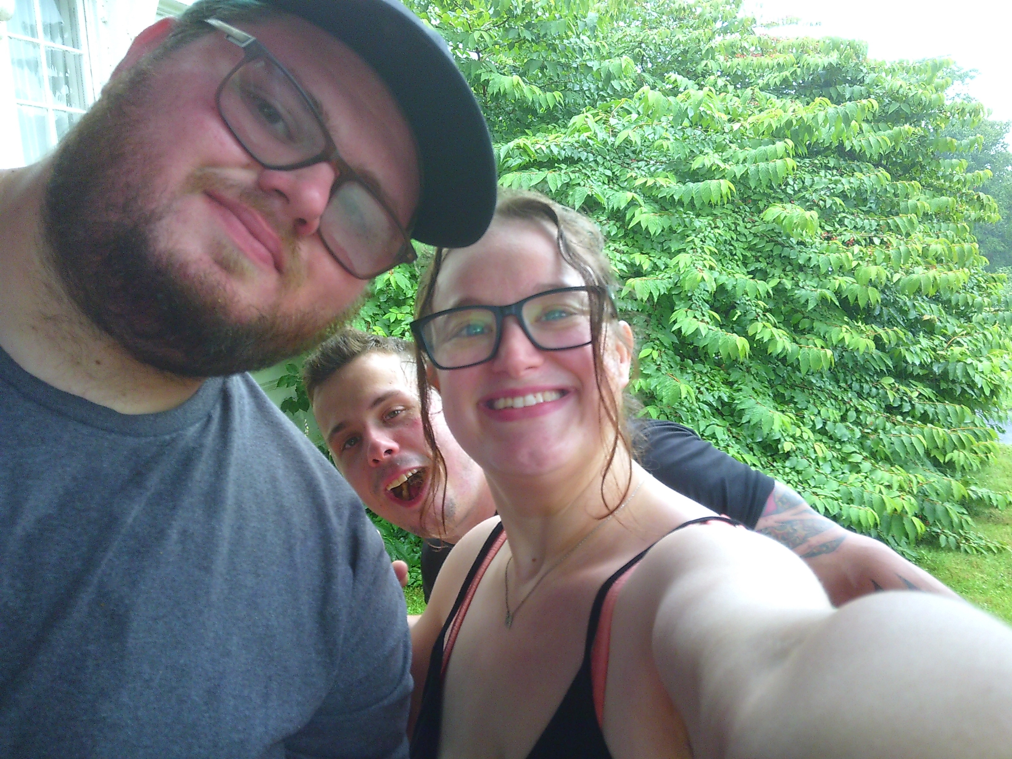 A selfie where a smiling woman is holding the camera, with a man in front of her and a man behind her. The man behind her is smiling with an open mouth