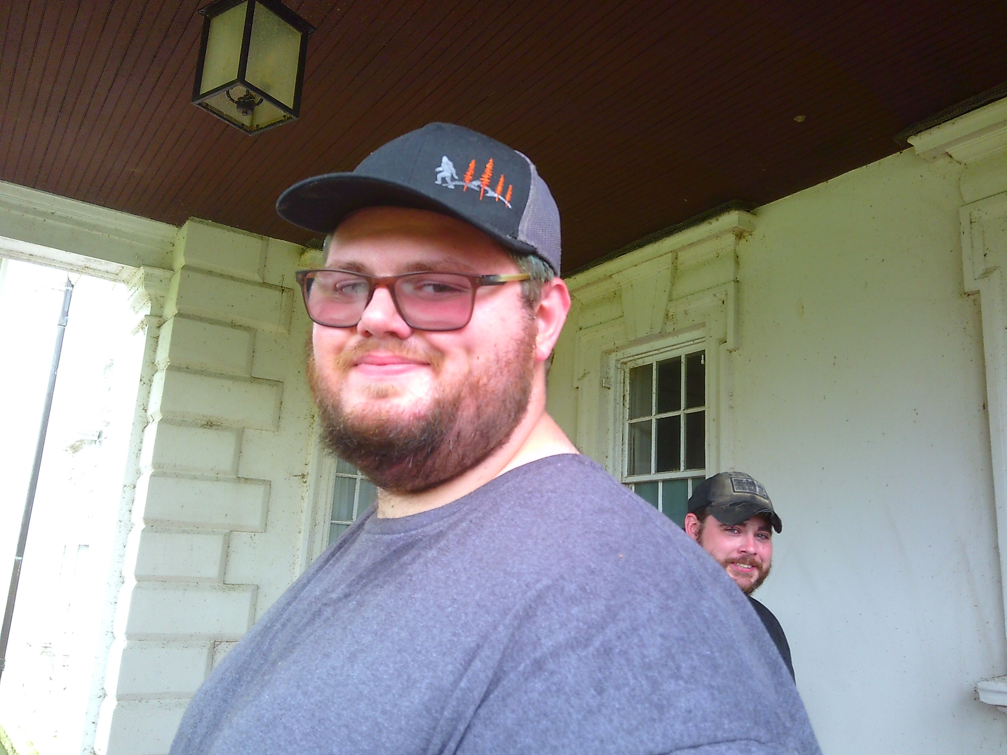 A photo of a man with a baseball cap and grey shirt on smiling towards the camera. Another man also looks at the camera from the background