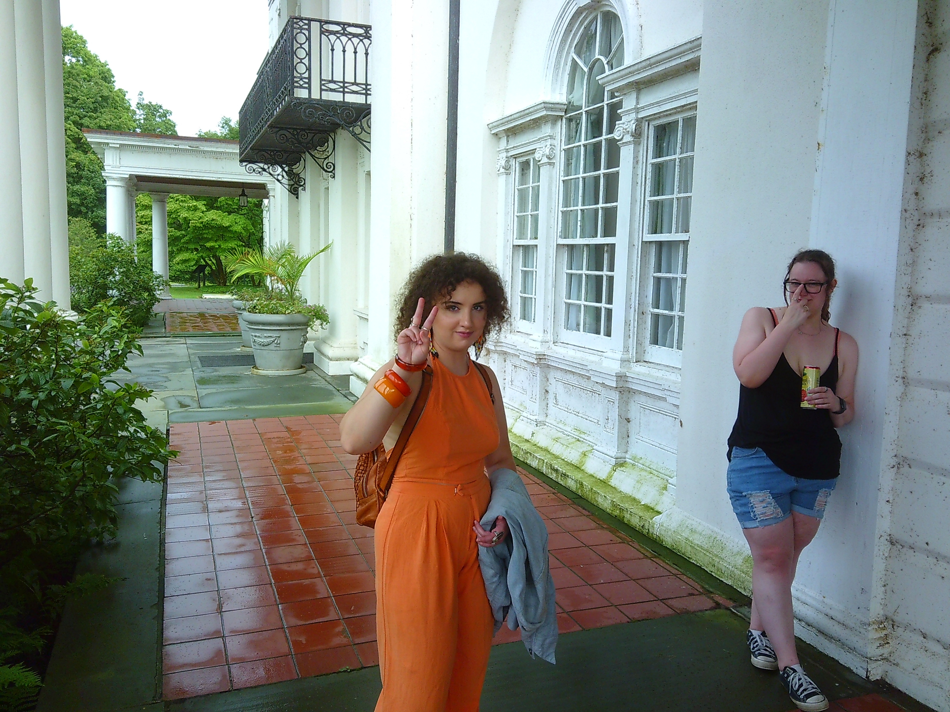 Photo of a woman in an orange outfit giving the peace sign. There is another woman adjusting her glasses towards the right of the frame.