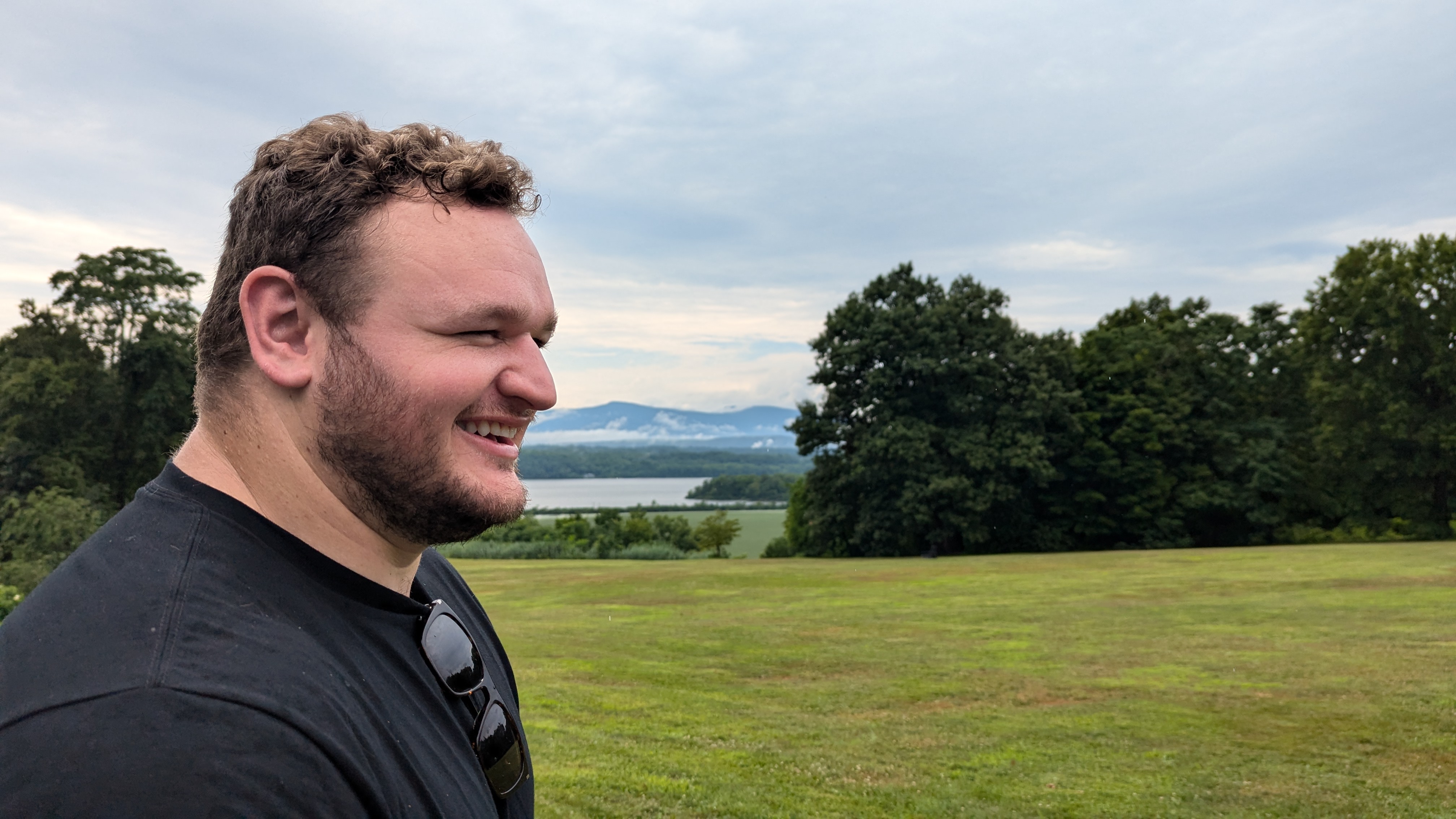 Photo of a man smiling in front of a green lawn with a river and mountain off in the distance