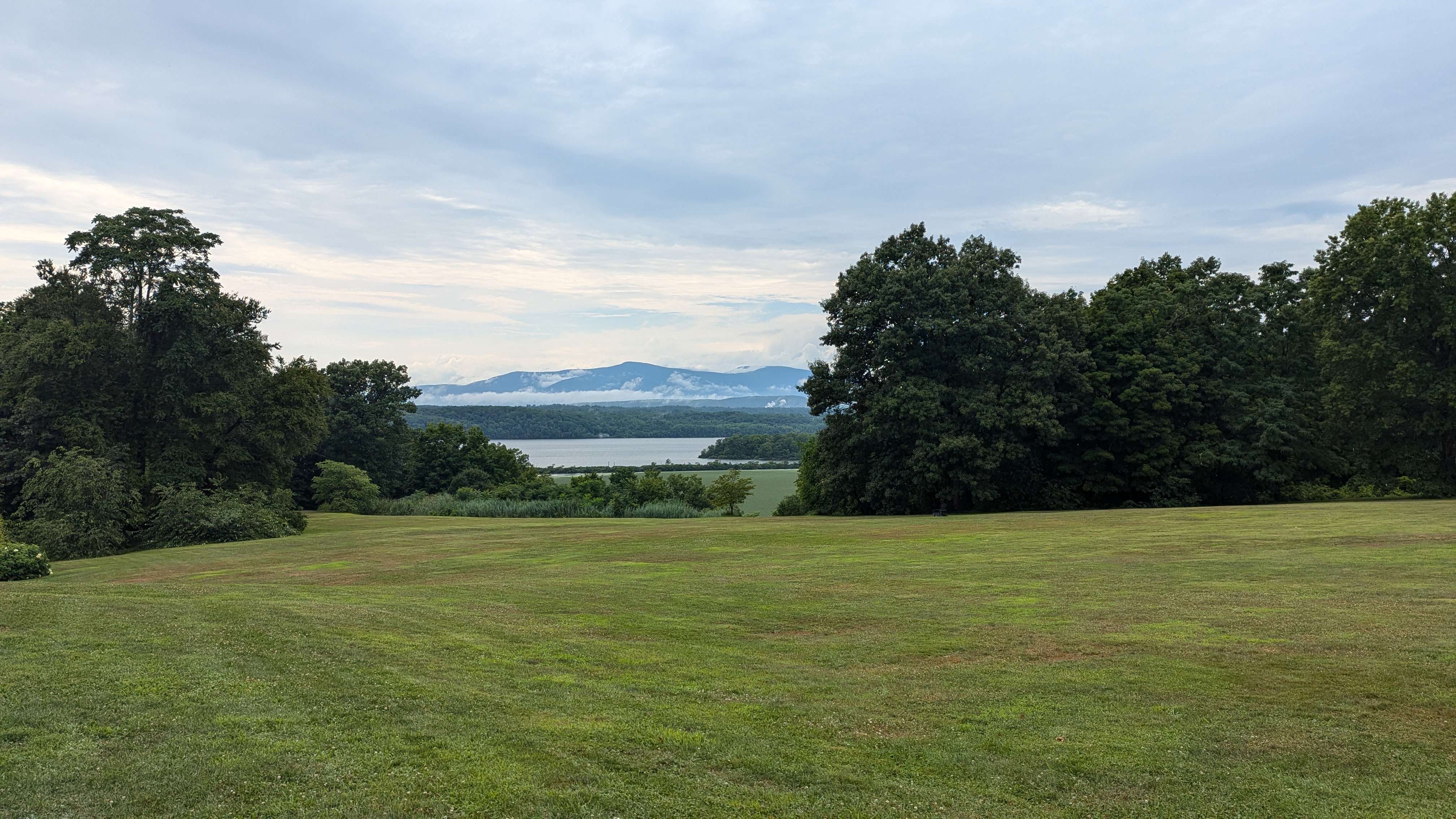 Photo of a green lawn with some trees, with a river in the mid ground and a mountain in the background