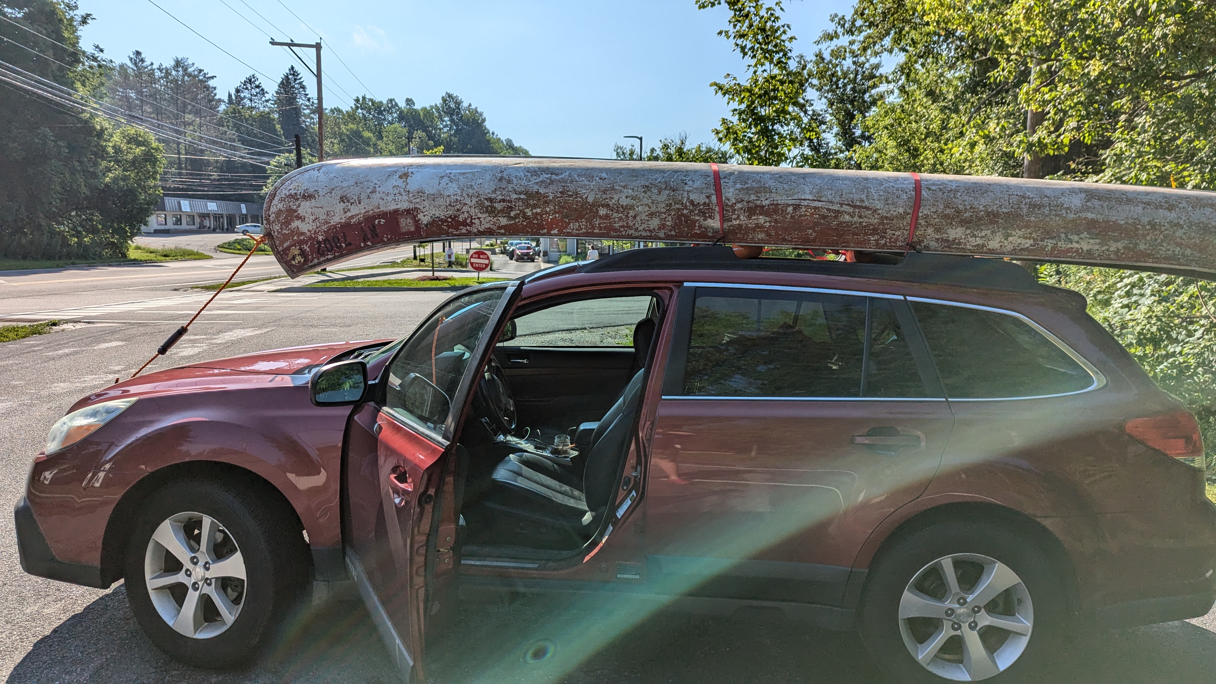 Red subaru outback station wagon with a metalic somewhat red canoe on top. There is an orange rope going from the front of the canoe to the front of the car, as well as two red cargo straps tying the canoe to the car.