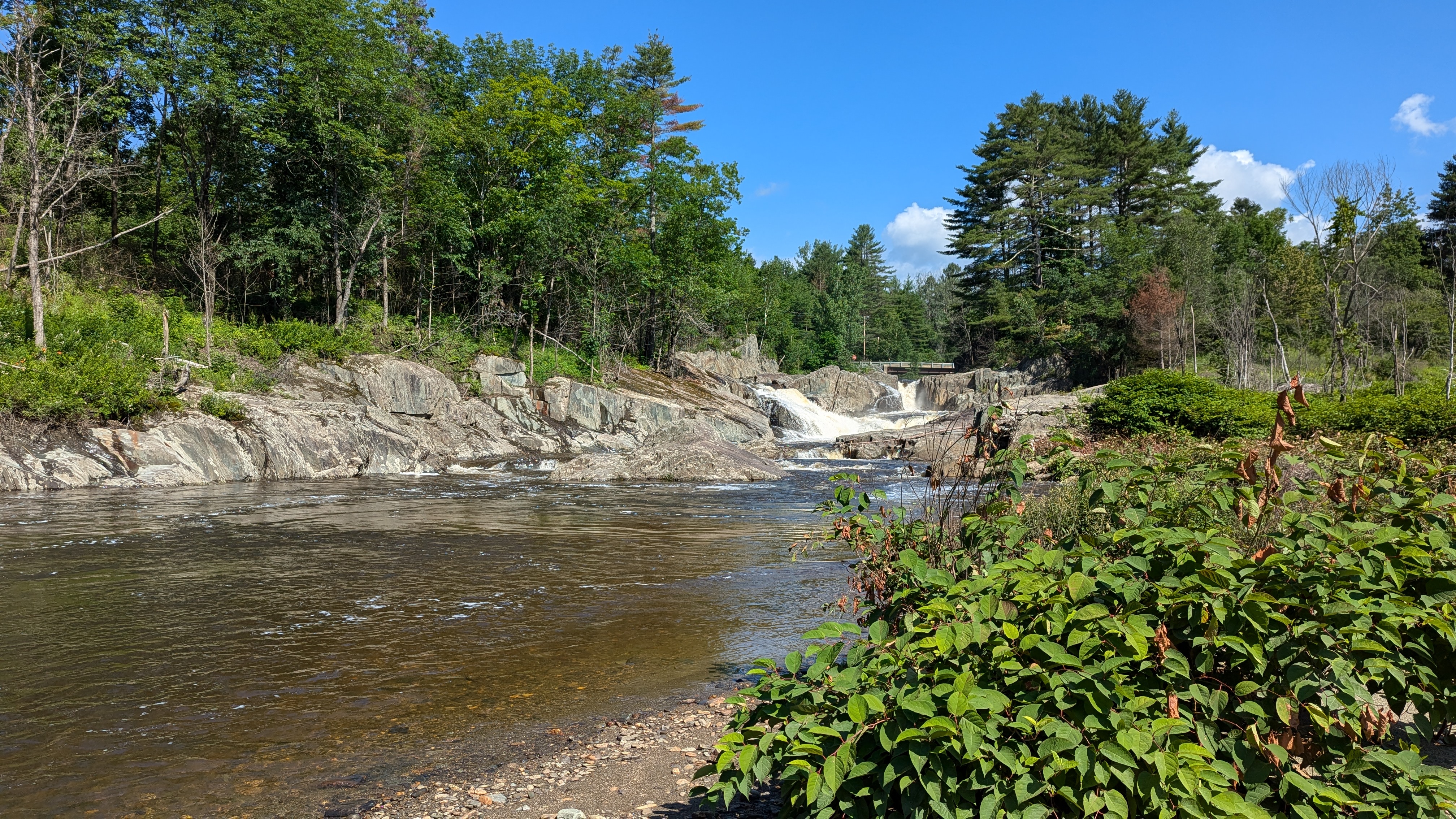 image of a waterfall in the distance with a bridge behind it. Leading up to the waterfall is the stream, with some vegetation on the right hand side ofthe photo. There are large rocks leading up to the waterfall.