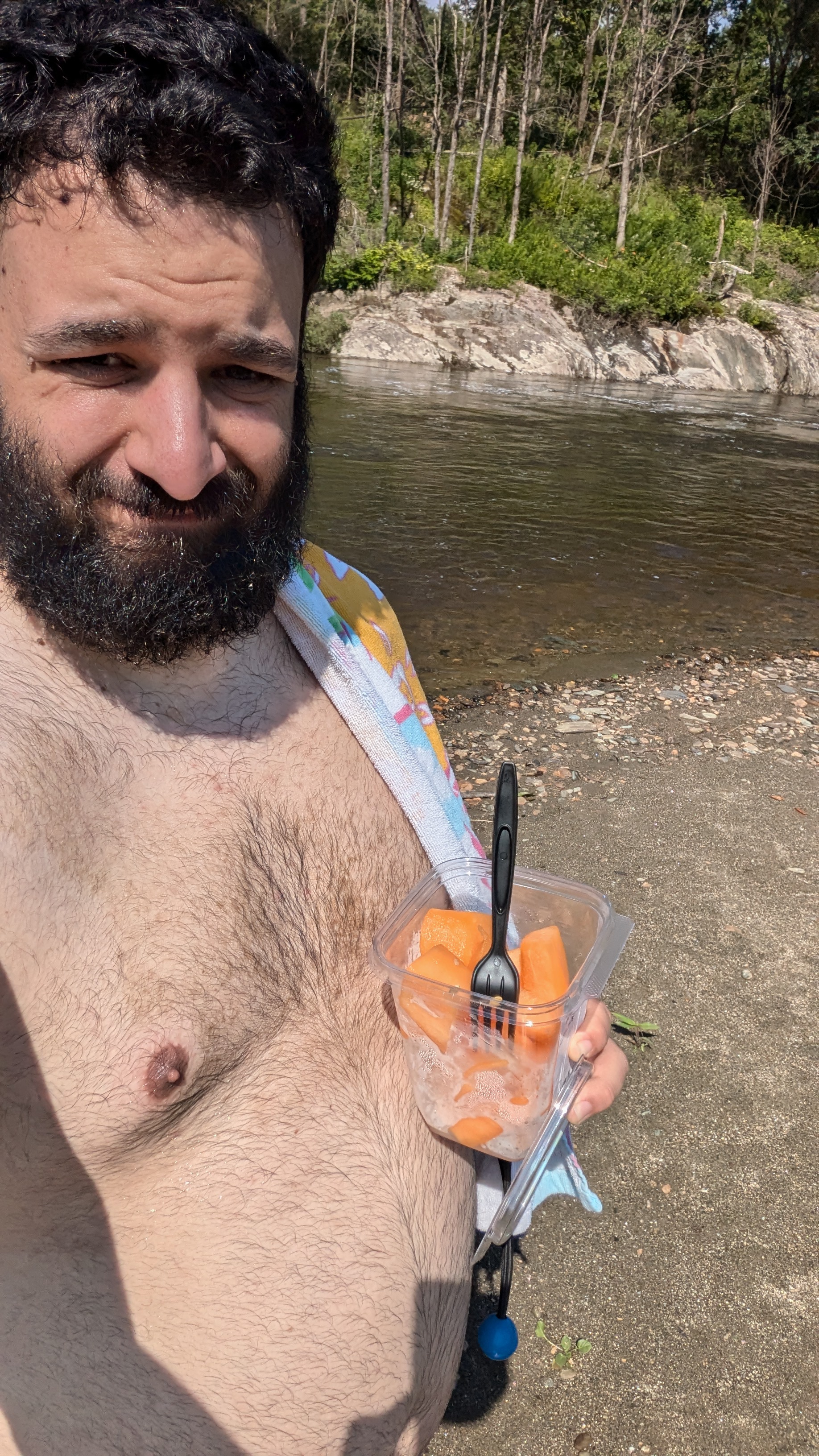 a selfie of a man with a dark beard and hair with a cup of cantelope in his hand. The man is shirtless and a bit heavyset. He is standing in front of what looks like a river.