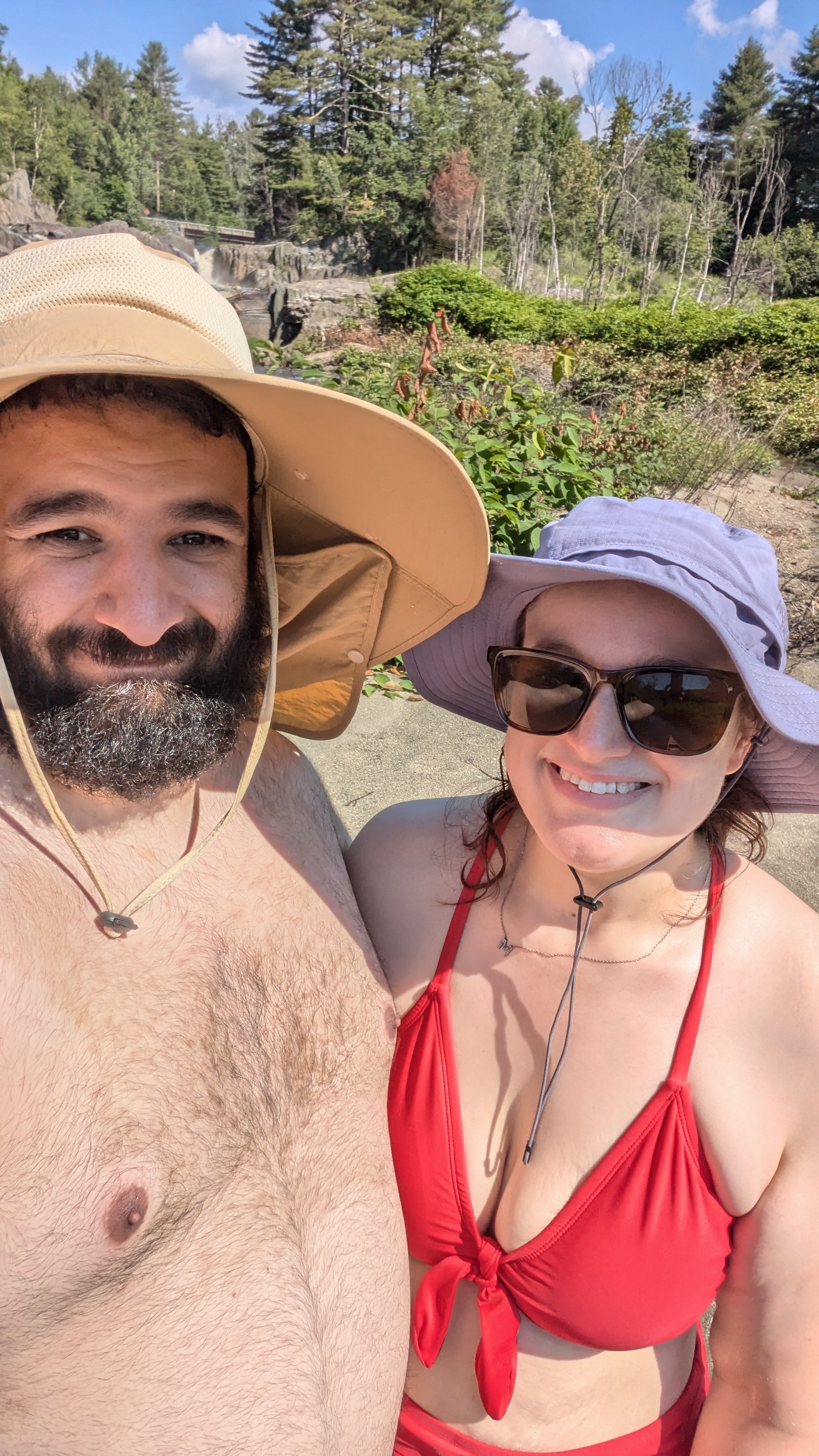 a photo of a man and woman standing next to each other taking a selfie. The man is shirtless with a wide brimmed tan hat, while the woman is in a red bikini with a purple wide brimmed hat.