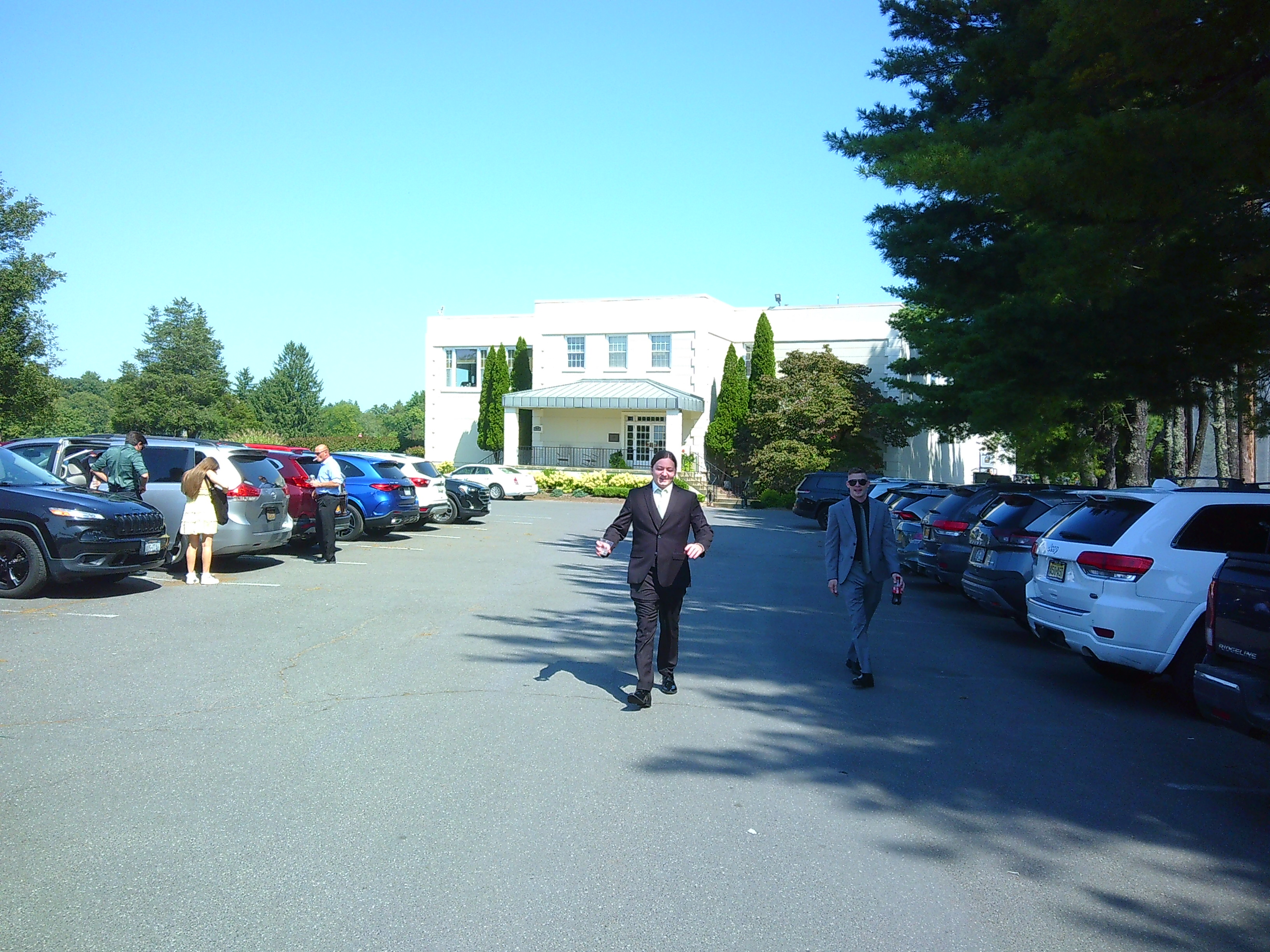 photo of a man (The groom, Sam) in a black tux walking towards the camera. He's outside in a parking lot, with a white building behind him.