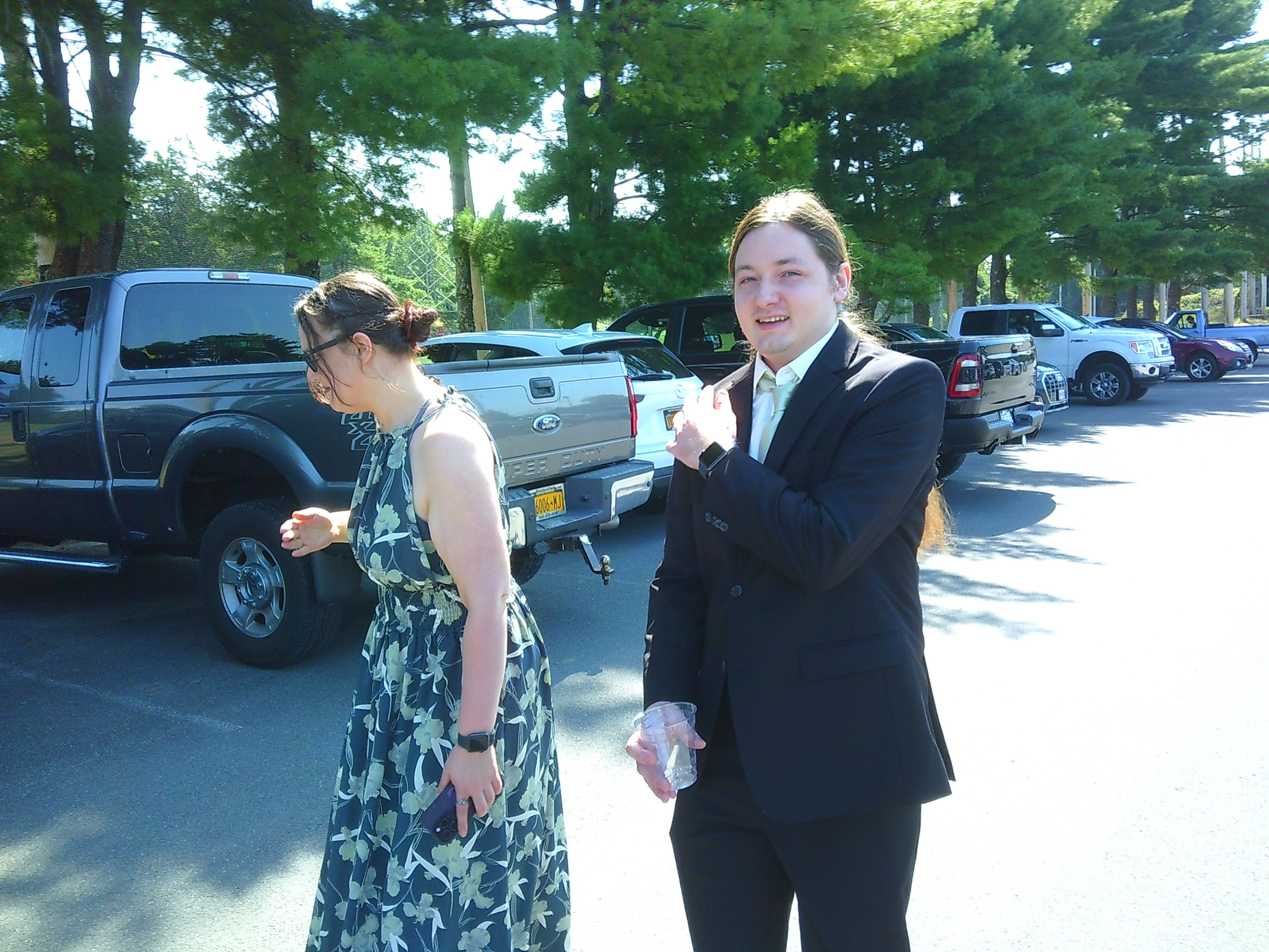 photo of a man (the groom, Sam) and a woman (Michelle) walking towards the camera, but the camera is off to the side a bit. The man is looking at the camera while brushing his soldier, while the woman is faced away from the photo. The man is in a black tux and the woman in a green flowy dress