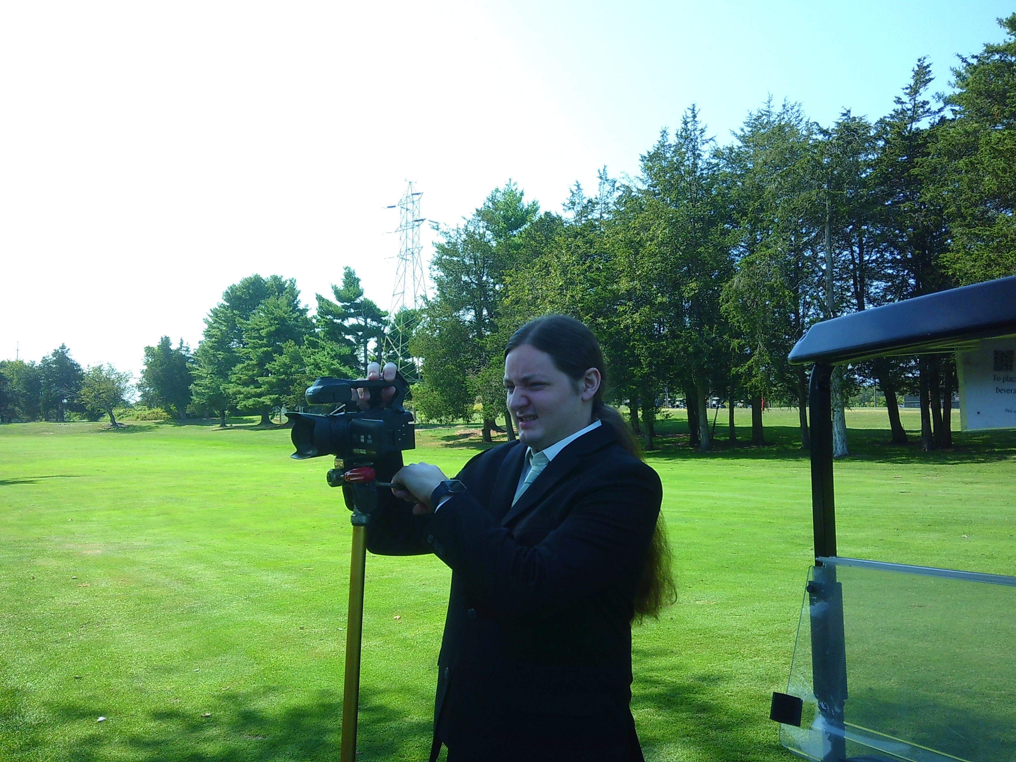 Photo of Sam setting up the camcorder. He is on a green golf field, with some trees in the background