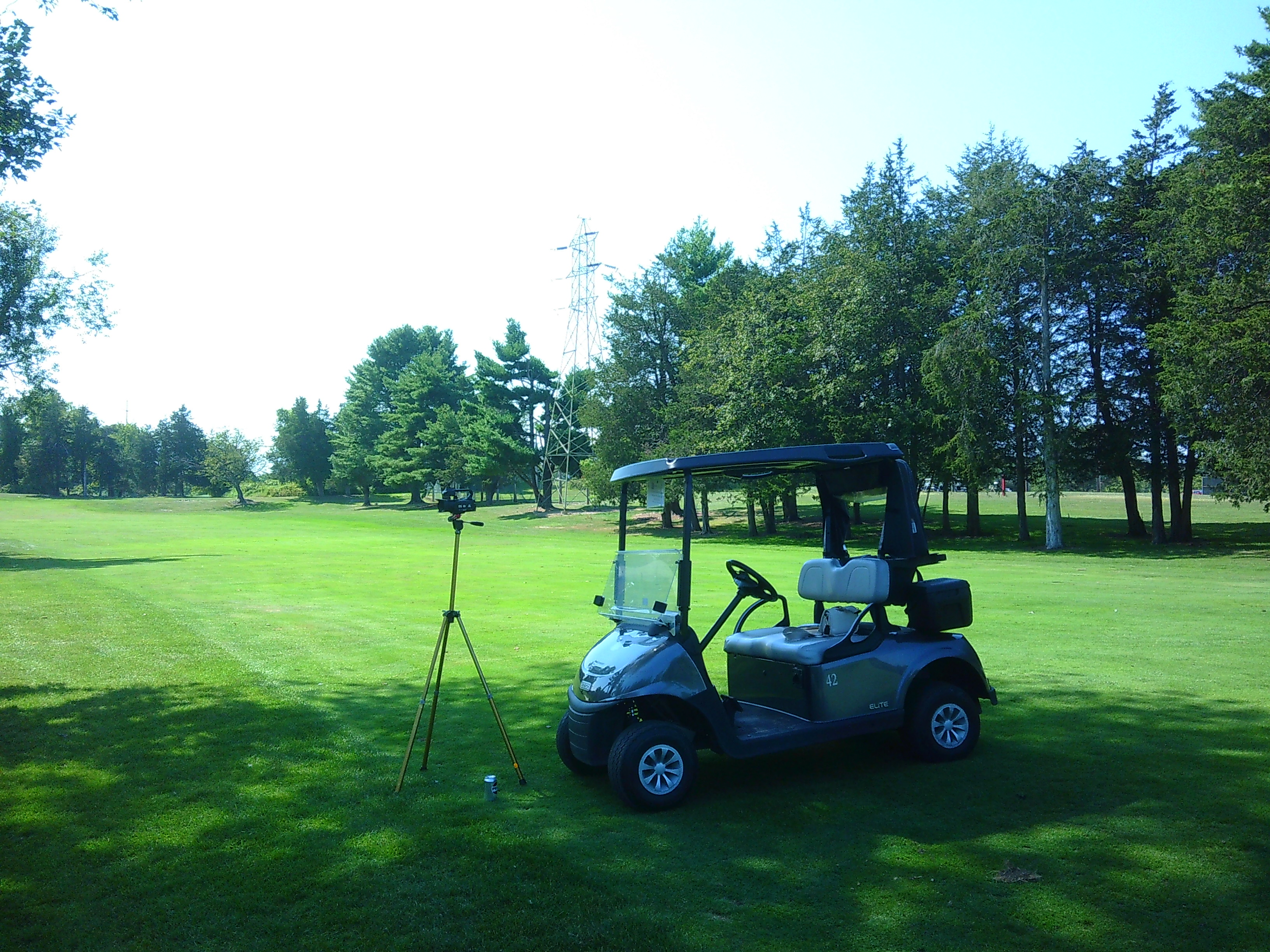 photo of a camcroder on a tripod on a green golf field with a golfcart next to it