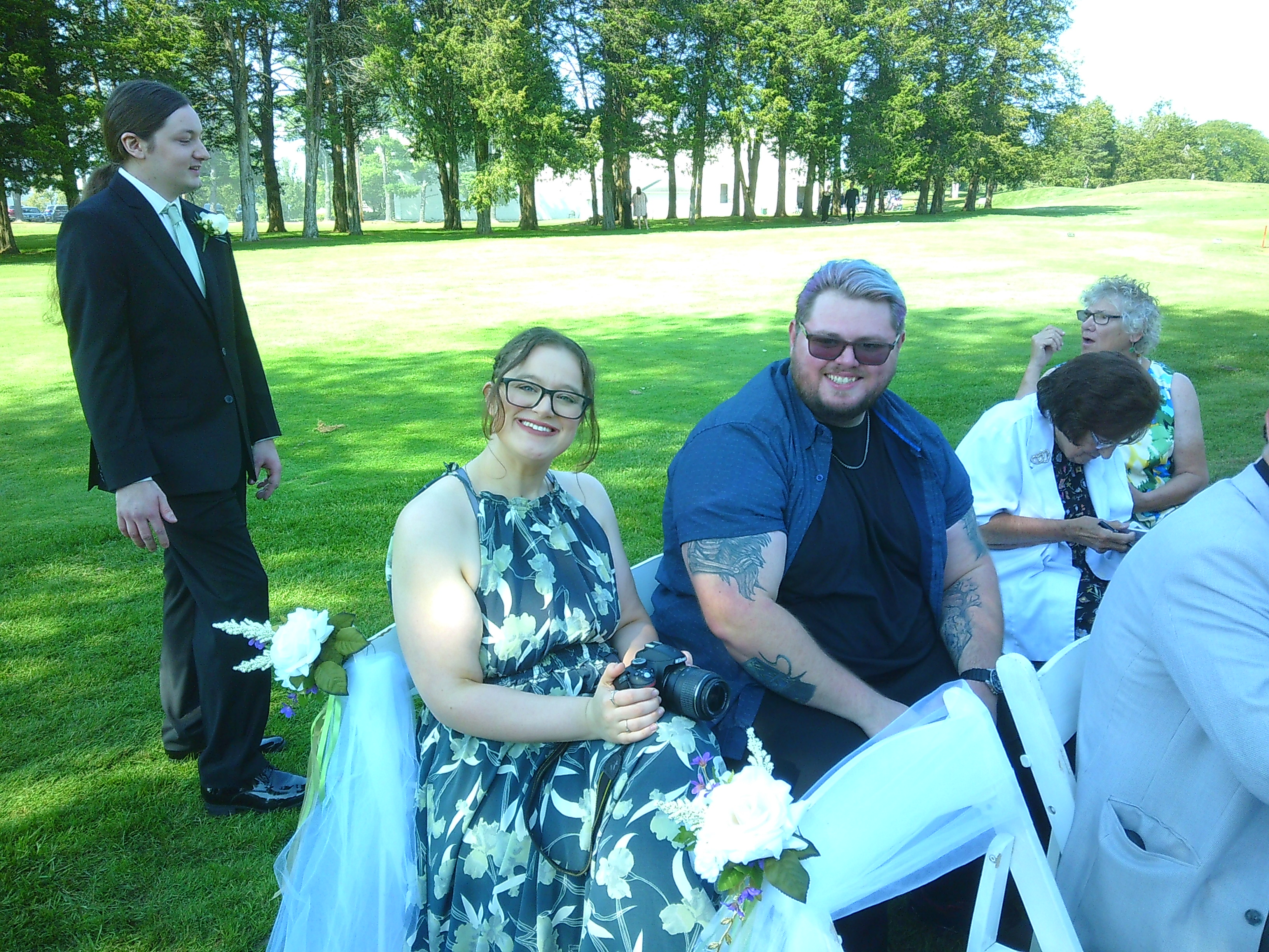 Photo of a woman (Michelle) and a man (Dane) sitting in white folding chairs on a golf field. The woman and man are both smiling at the camera. The woman is holding a Nikon d3200 digital camera