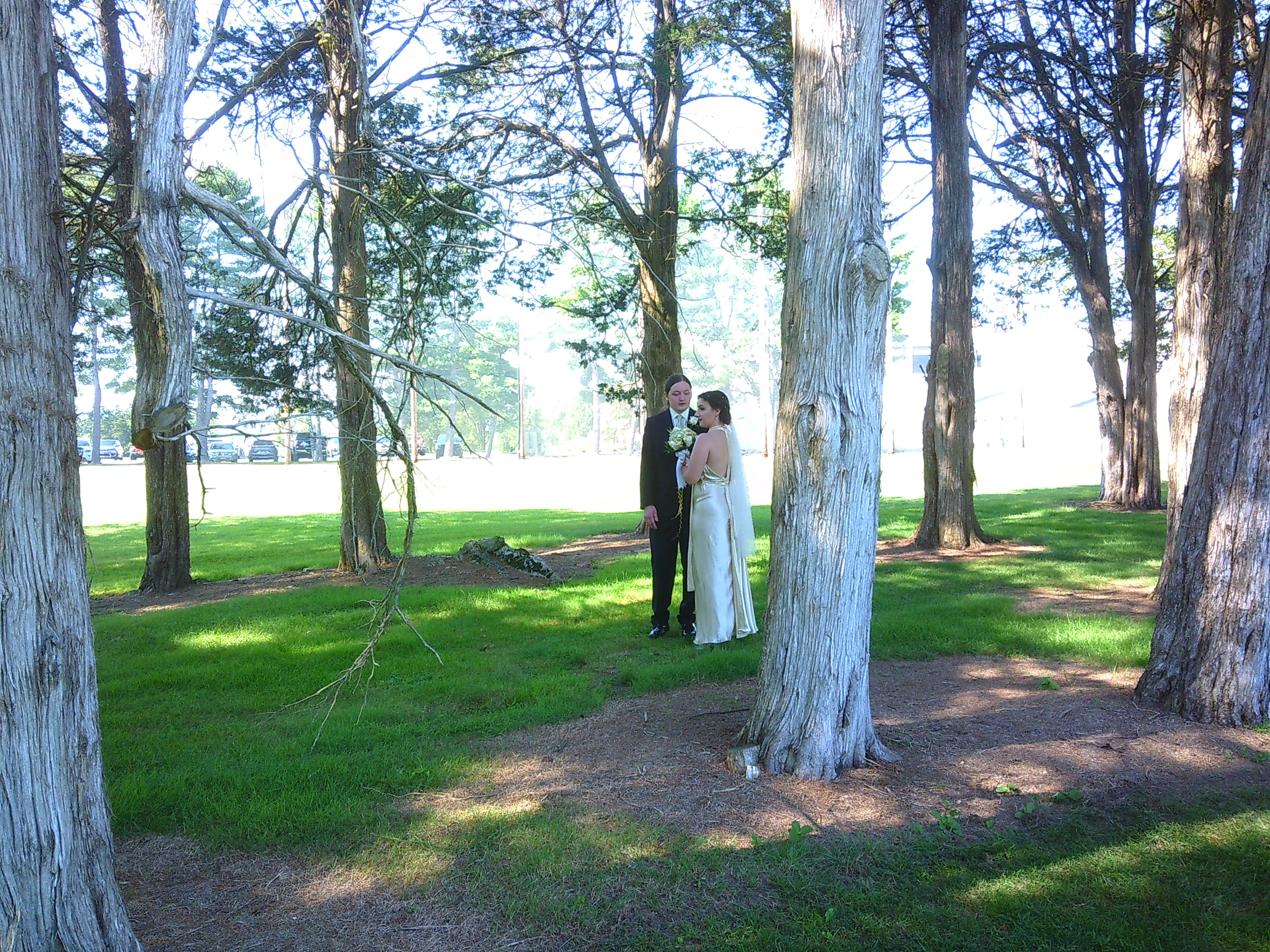 Photo of a man (The Groom, Sam) and a woman (The Bride, Zakiah) standing in between some rows of trees on a green spot of trash. The woman is holding a bouquet  of flowers and the man is looking at the woman. The woman is an off white dress with a veil hanging behind her head.