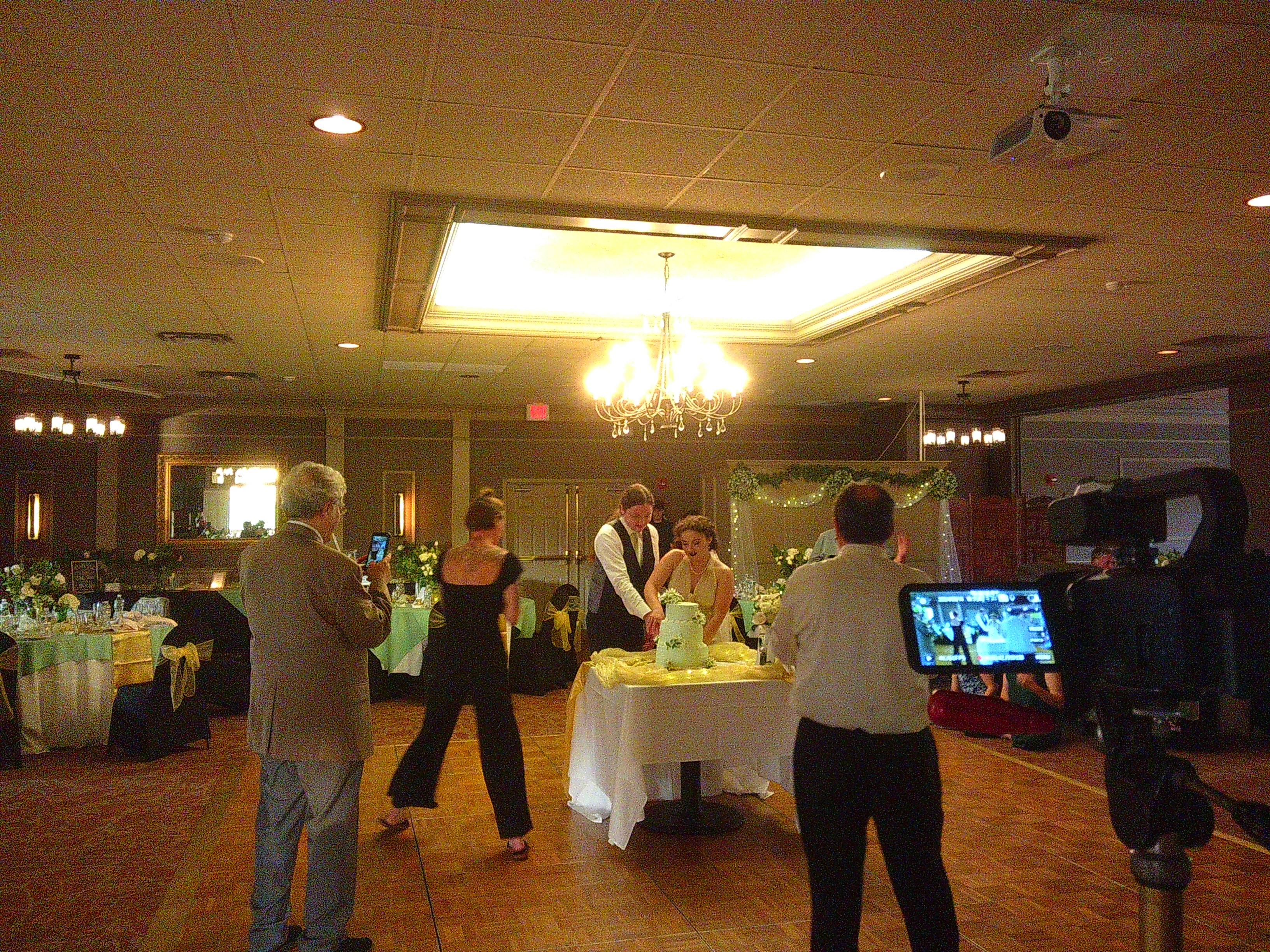 Photo of the bride and groom, Zakiah and Sam, cutting their wedding cake. It's a green cake and they're cutting it on a table in the middle of the dance floor. Zakiah has her hand over Sams as they cut into the cake.