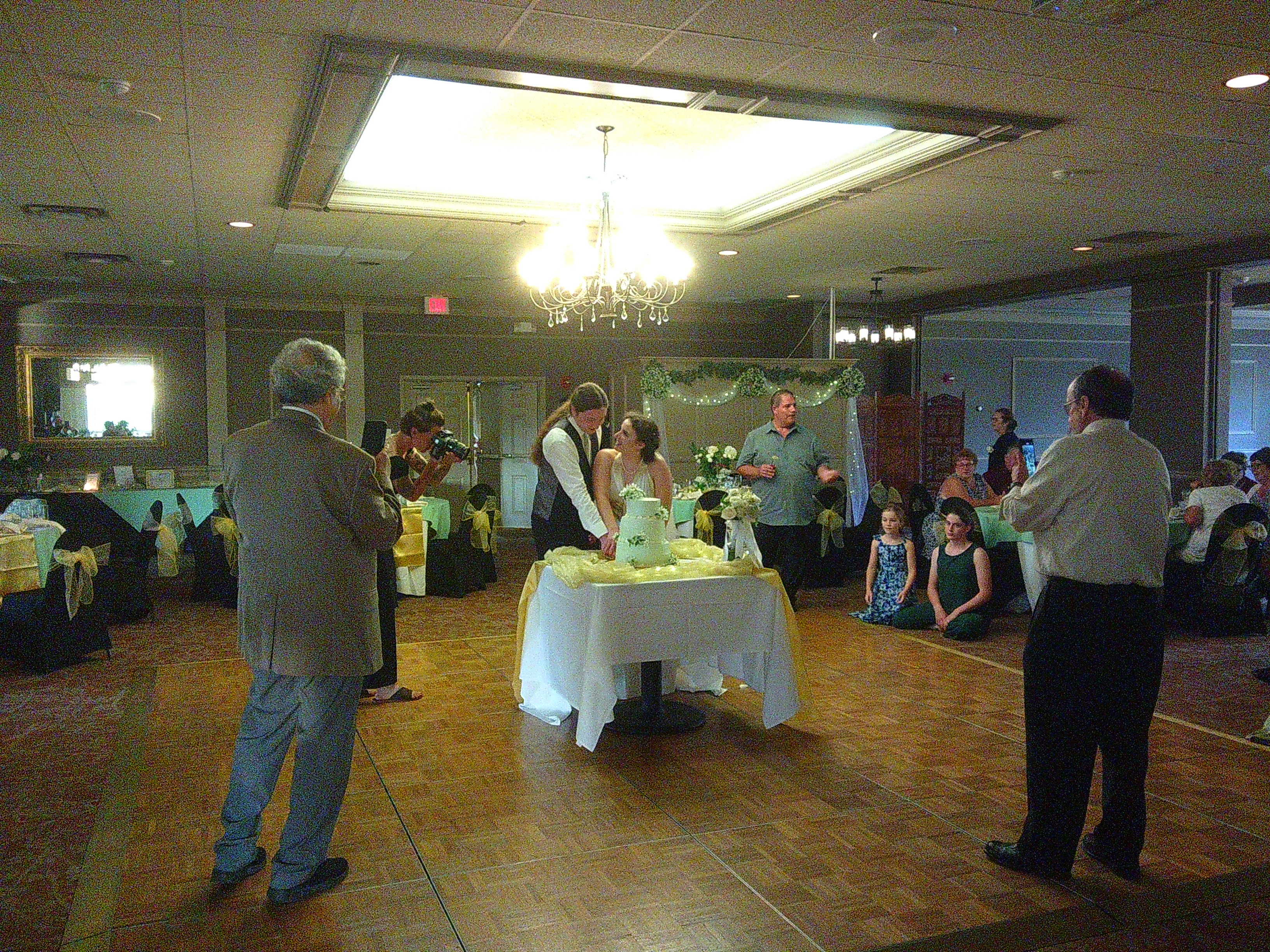 Photo of Groom Sam and Bride Zakiah cutting their cake in the center of a dance floor. Zakiah is looking up at Sam while Sam is looking at the knife as they cut into a Green cake.