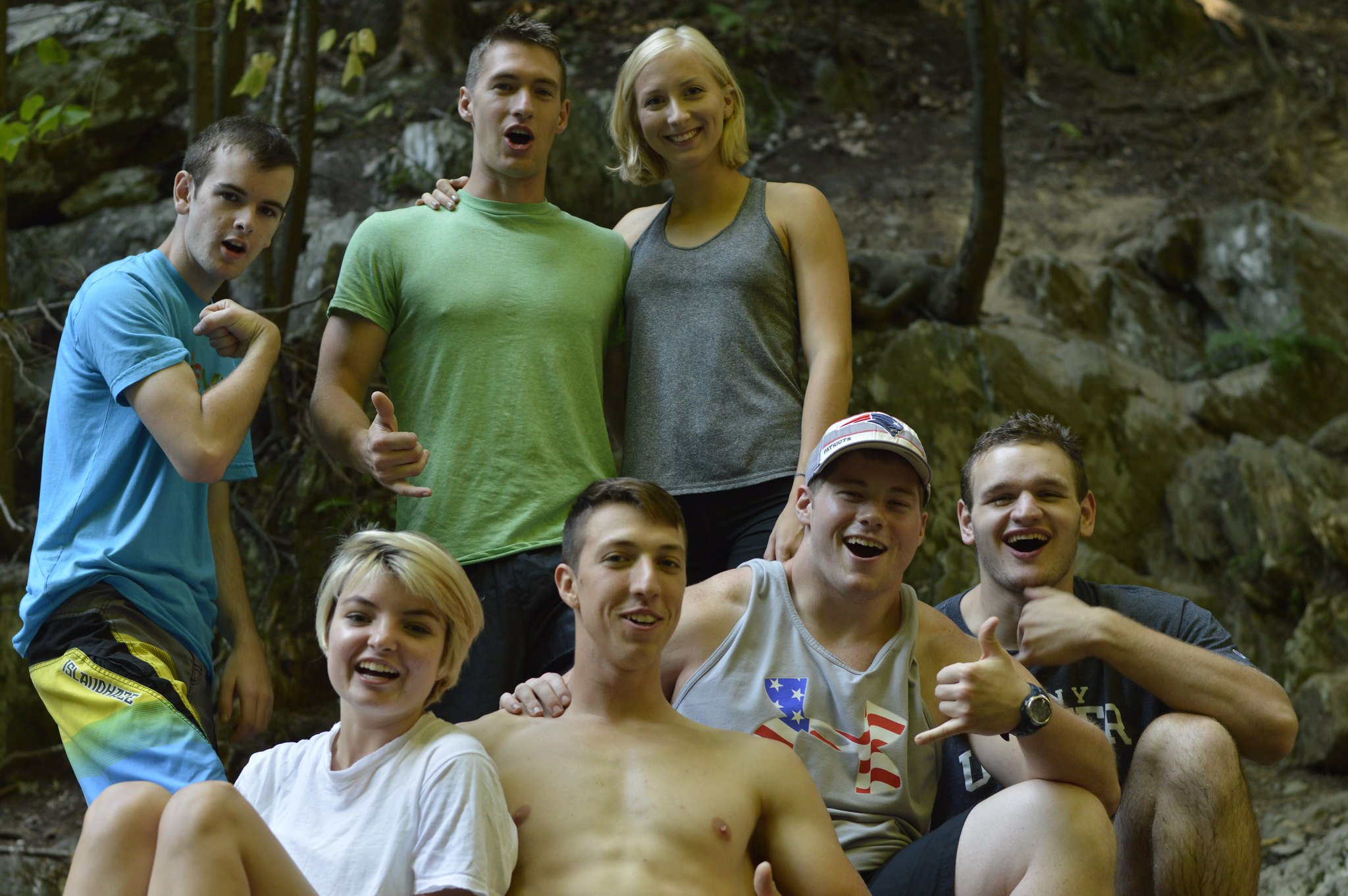 Photo of 7 people smiling for a photo. They're in the woods, with 4 of them sitting down and 3 of them standing up