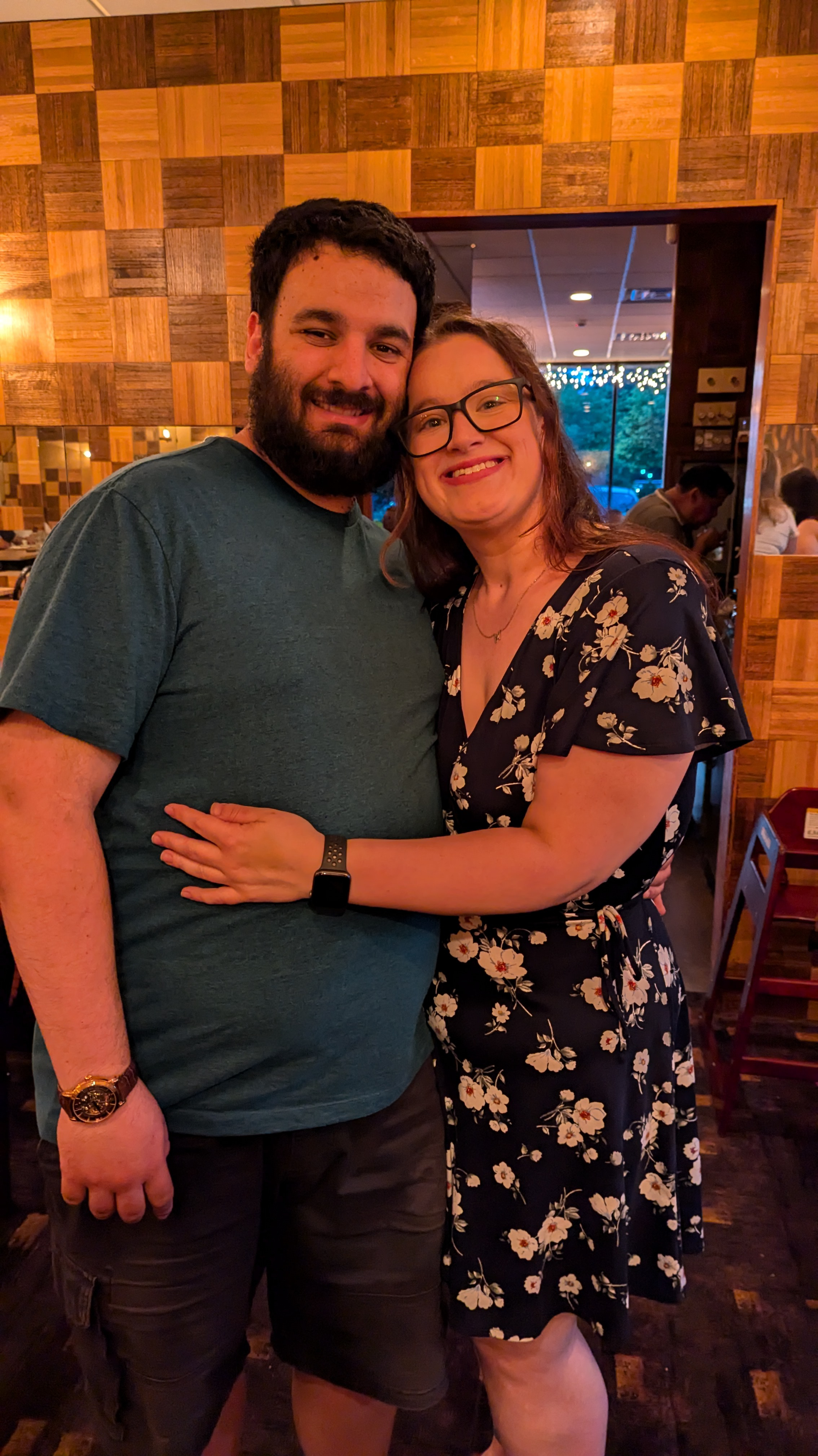 photo of a man faceing towards the camera smiling while a woman hugs the man and also faces the camera smiling