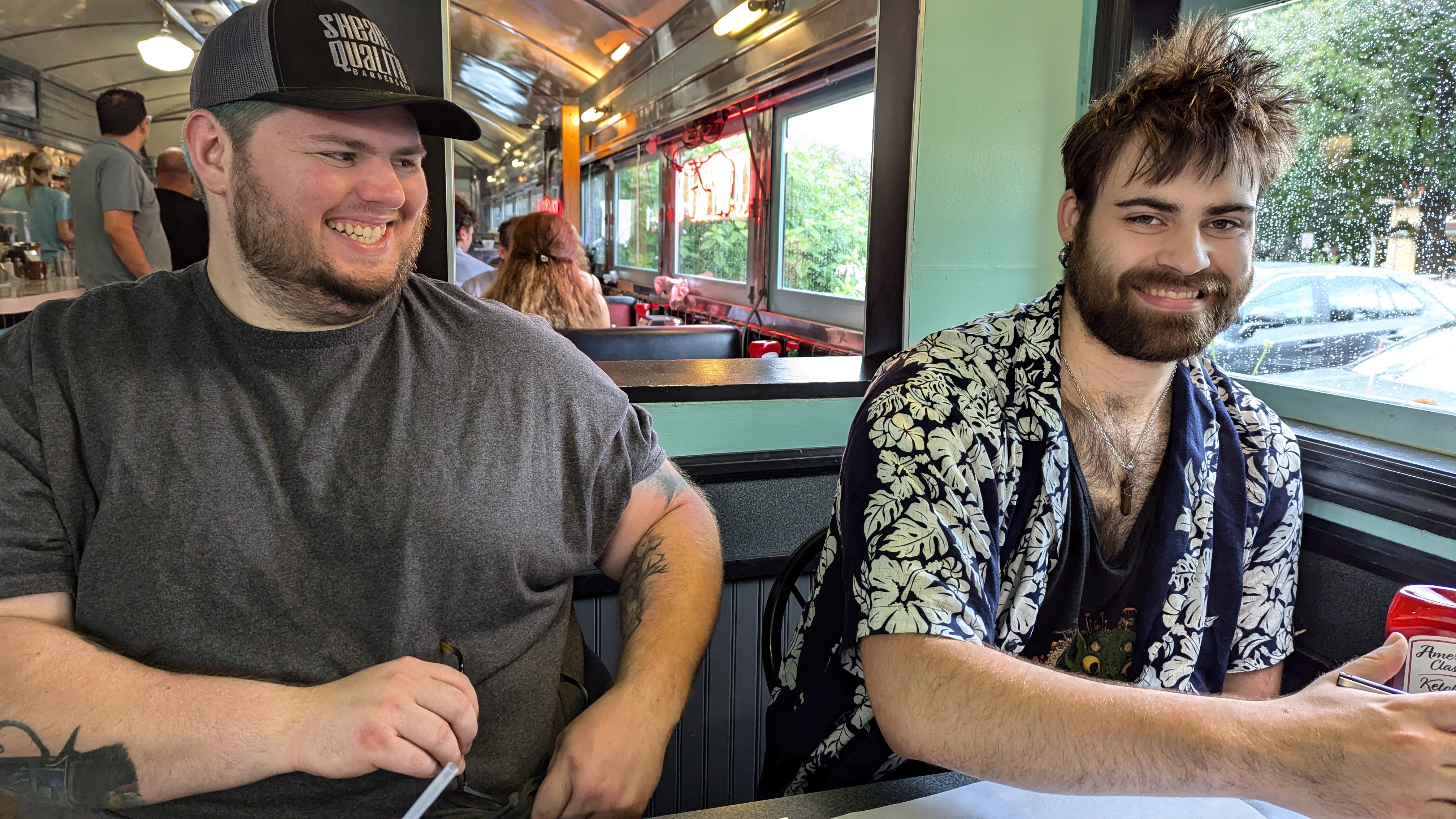 Photo of two men sitting next to each other at a diner booth. One is built like a brick house, the other more lean