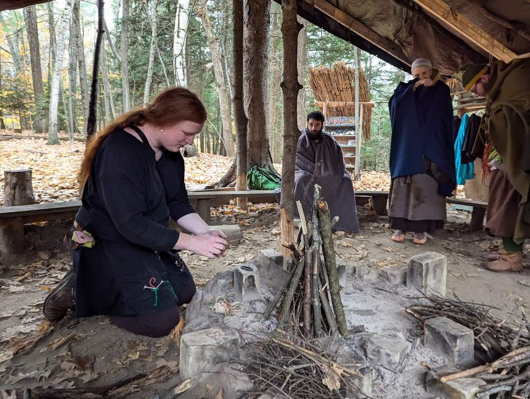 Photo of someone putting together a fire, with Steven wrapped in a grey blanket in the background looking towards the person putting the fire together.