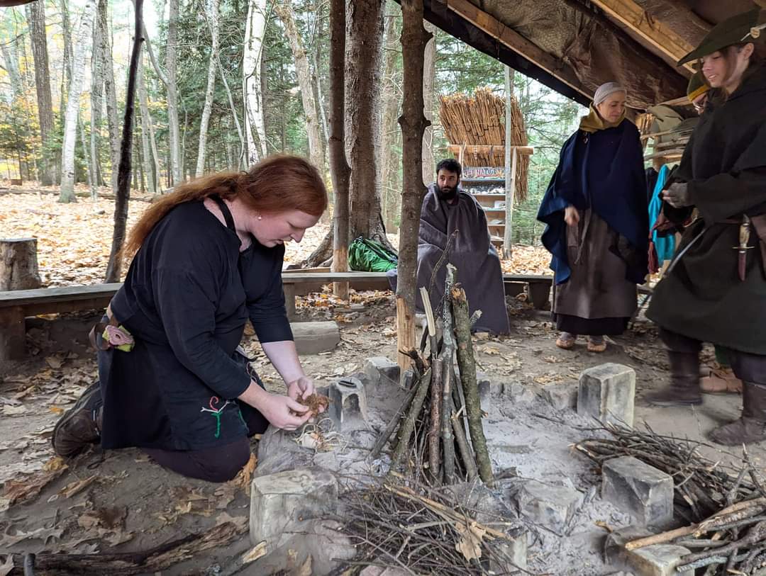 Photo of someone putting together a fire, with Steven wrapped in a grey blanket in the background looking towards the person putting the fire together.