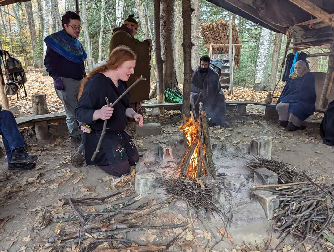 Photo of someone taking care of a lit fire, with Steven wrapped in a grey blanket in the background looking towards the fire.