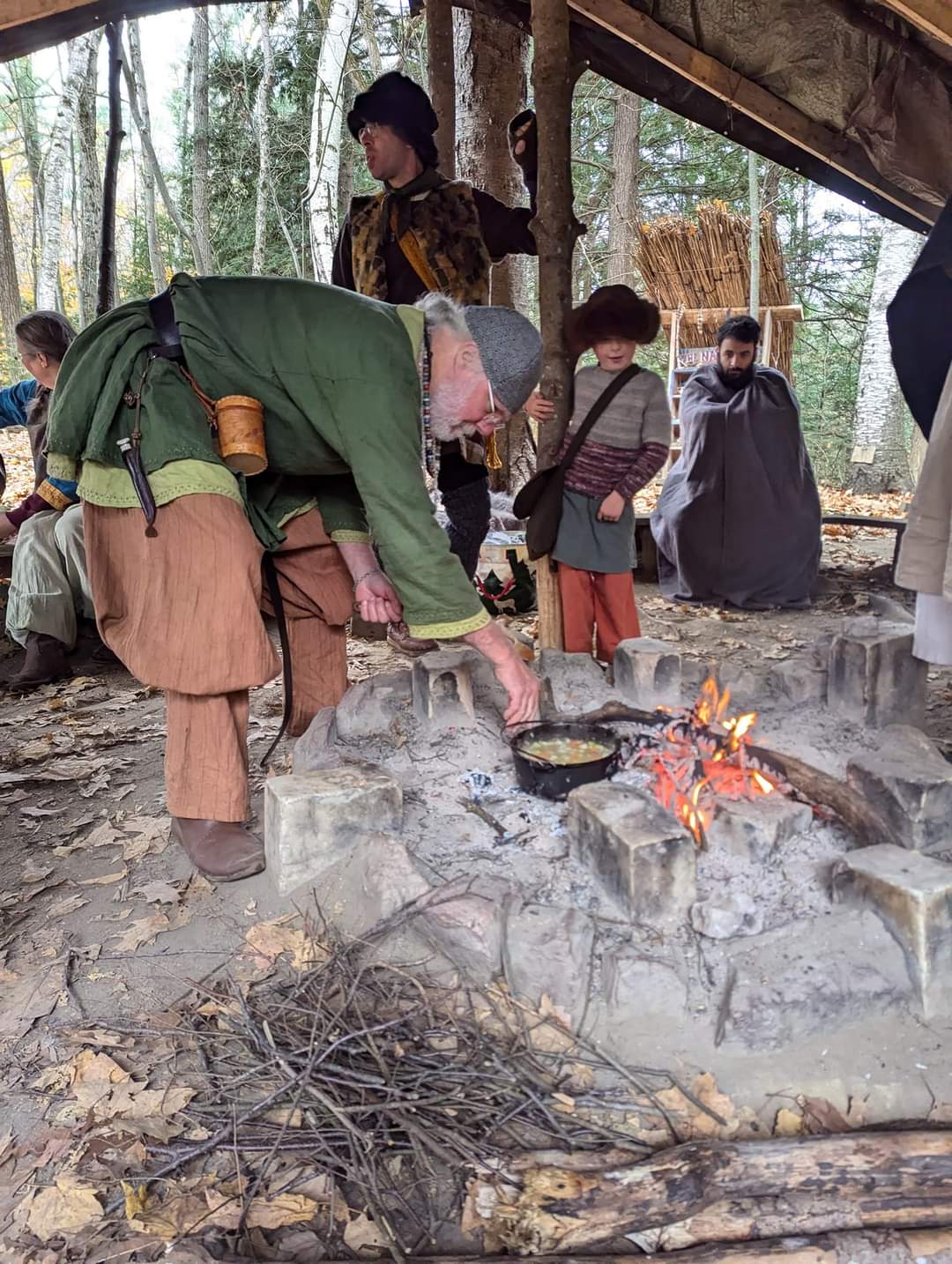 Photo of a man cooking a stew over the fire, with Steven wrapped up in a blanket looking at the fire in the background