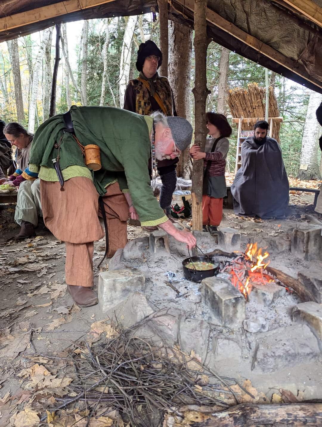 Photo of a man cooking a stew over the fire, with Steven wrapped up in a blanket looking at the fire in the background