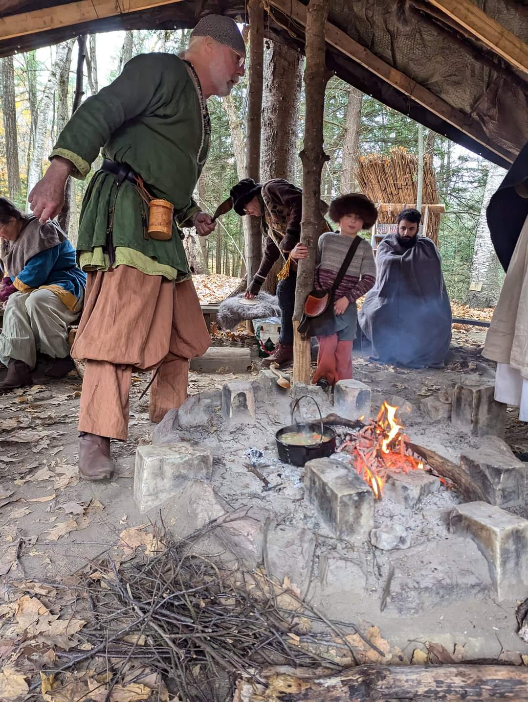 Photo of a man cooking a stew over the fire, with Steven wrapped up in a blanket looking at the fire in the background