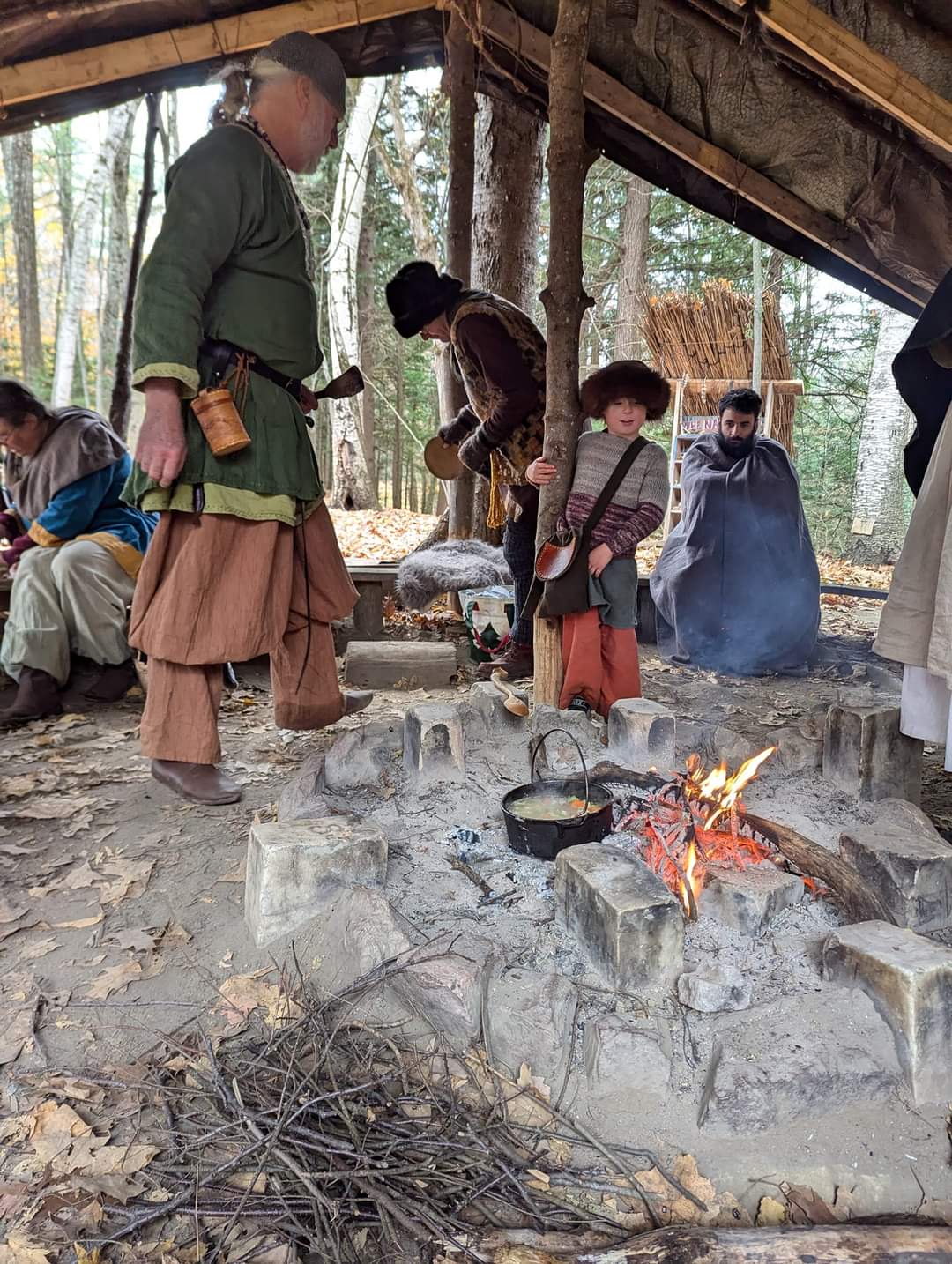 Photo of a man cooking a stew over the fire, with Steven wrapped up in a blanket looking at the fire in the background