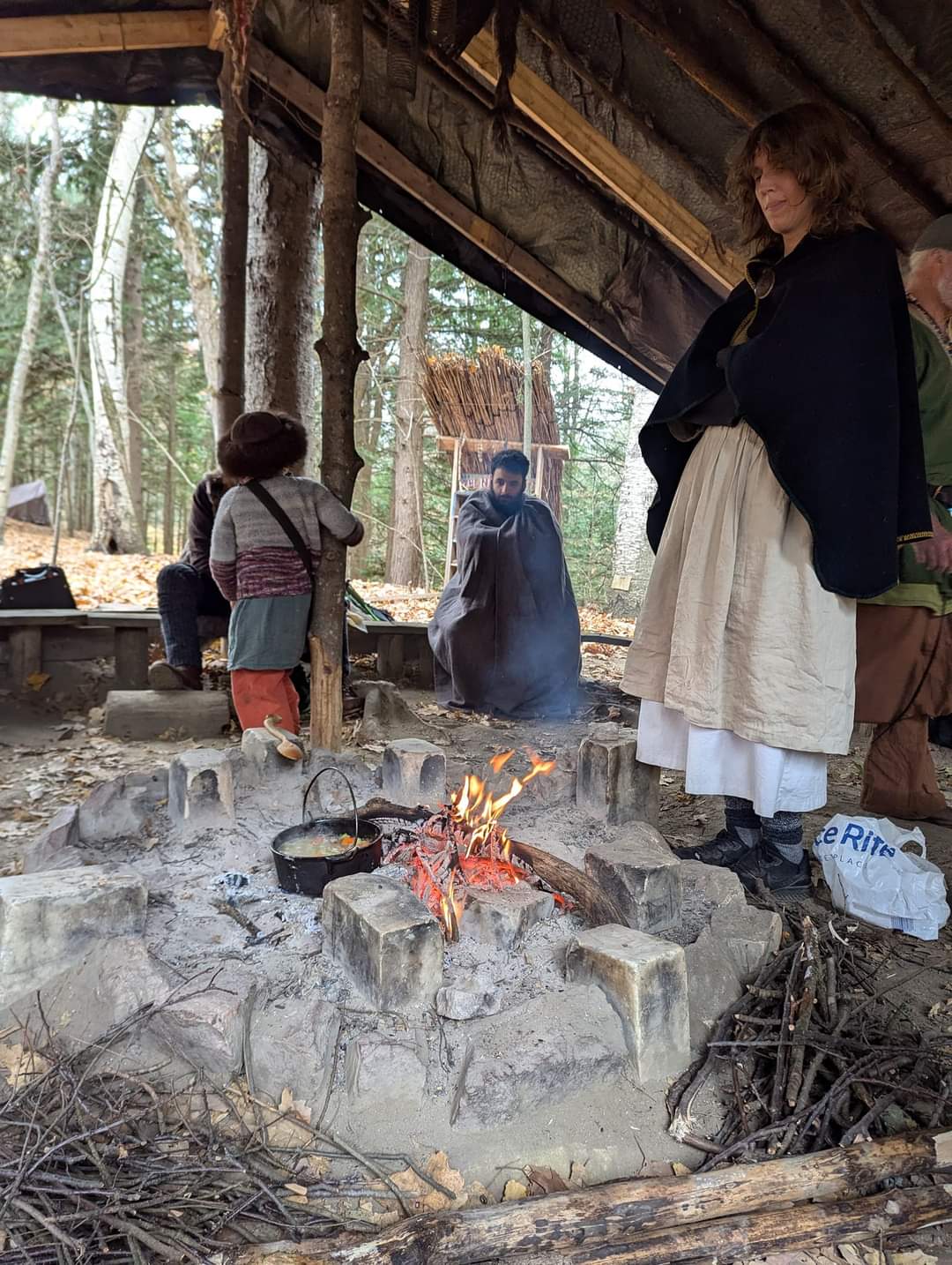 Photo of a person standing around the fire, looking at it, with Steven wrapped up in a blanket looking at the fire in the background