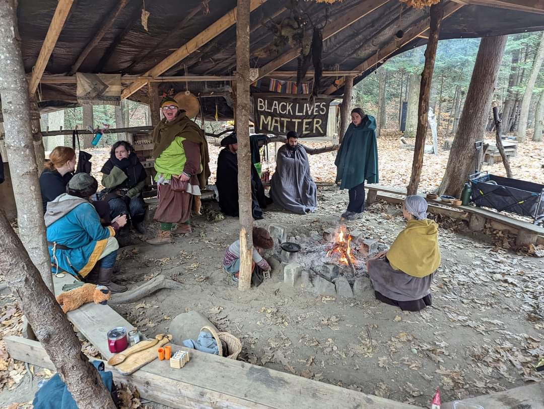 Photo of a group of people sitting around a fire, with Steven wrapped up in a blanket in a different spot in the background