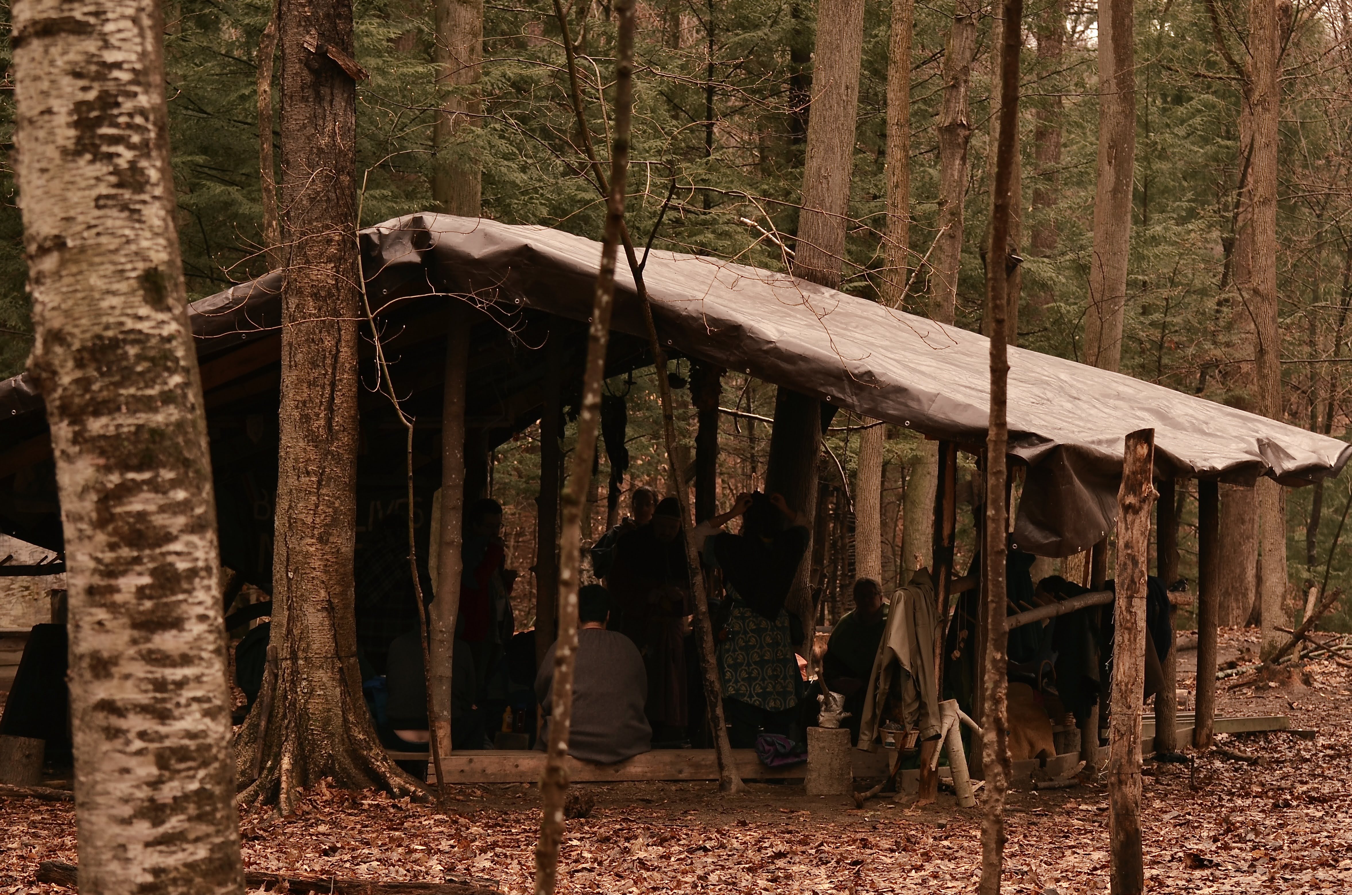 Photo of a shelter in the woods with a bunch of people inside of it. There is one large tarp, and no walls. There are benches along the outside, where the floor is indented a bit so the benches are not right on the bottom.
