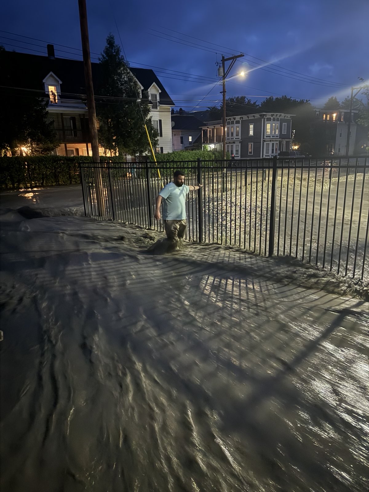 A night time photo of water flooding a driveway and surrounding streets with a man leaning on a gate while wading through the water