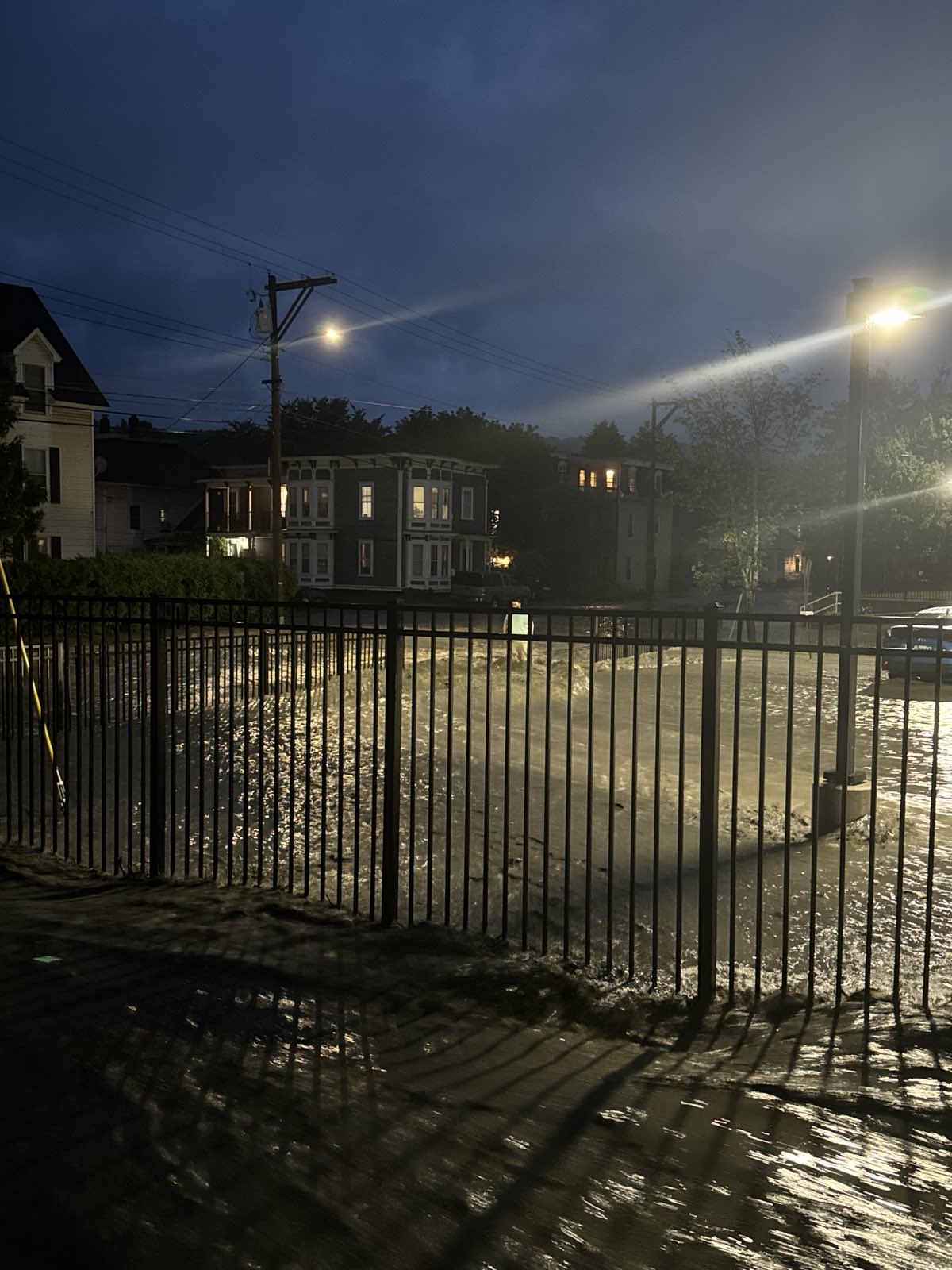 A night time photo of water rushing through a street and past a fence and parking lot, with a man in the distance trying to wade through the water