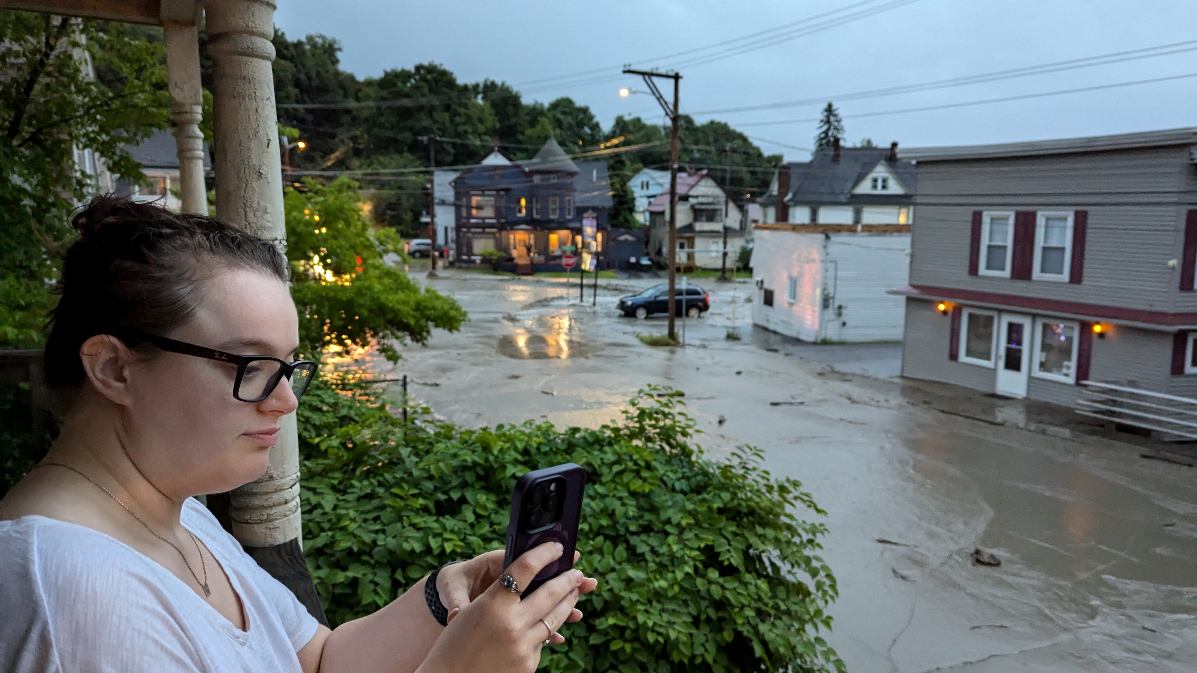 A woman can be seen taking a photo with her phone, which is pointed off camera. In the rest of the photo, you can see a lot of mud and flooded streets in a city area.