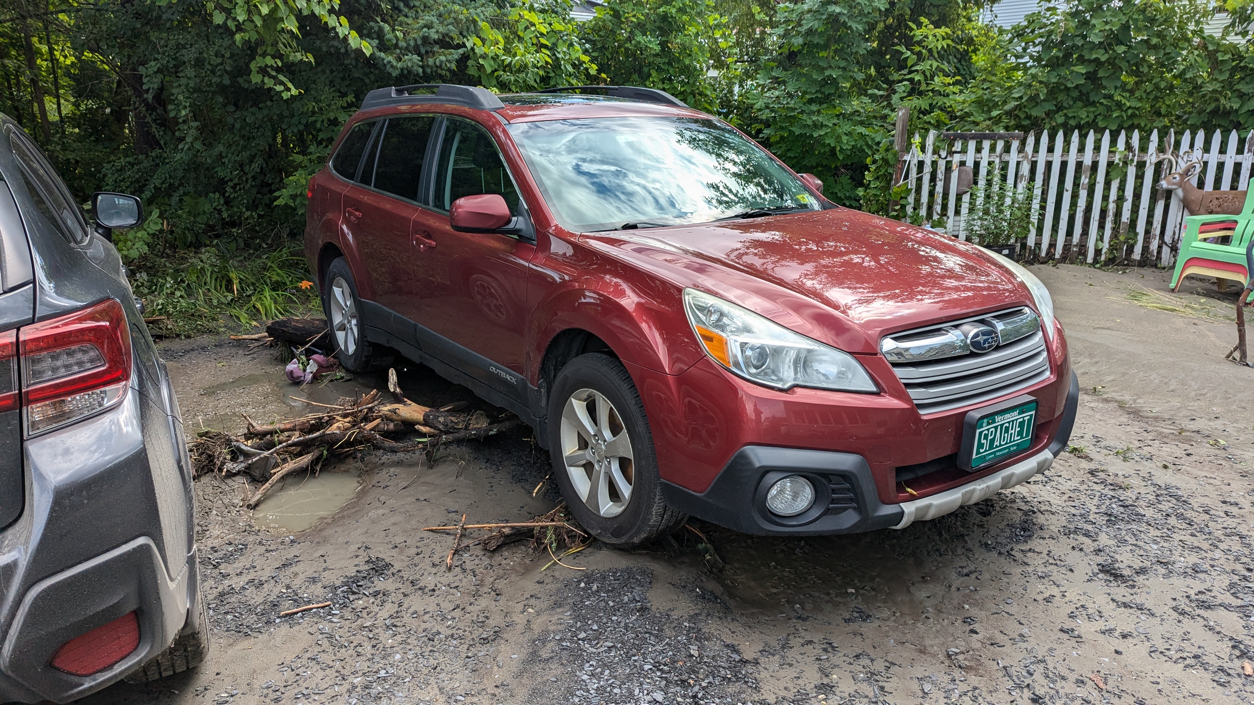 A red Subary Outback on a dirt driveway with a lot of wood sticking out from underneath it, and a lot of the dirt washed away from under the tires