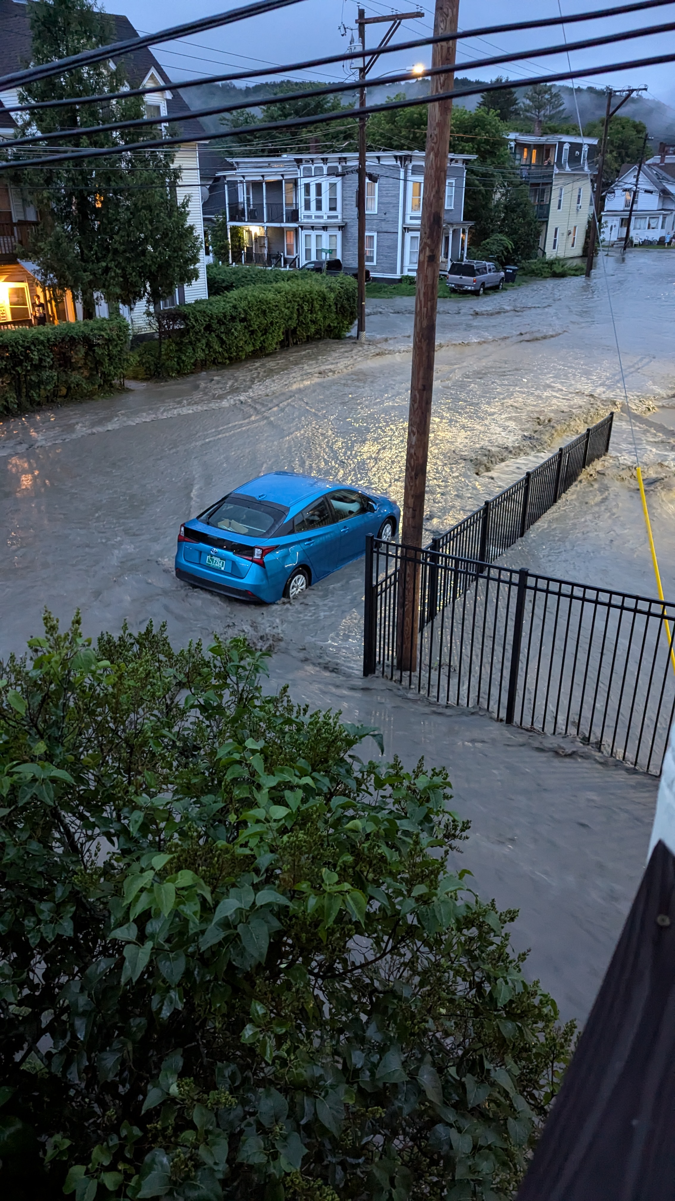 A photo of a flooded city street, with a bright blue Prius in it. The water is almost to the bottom of the prius doors