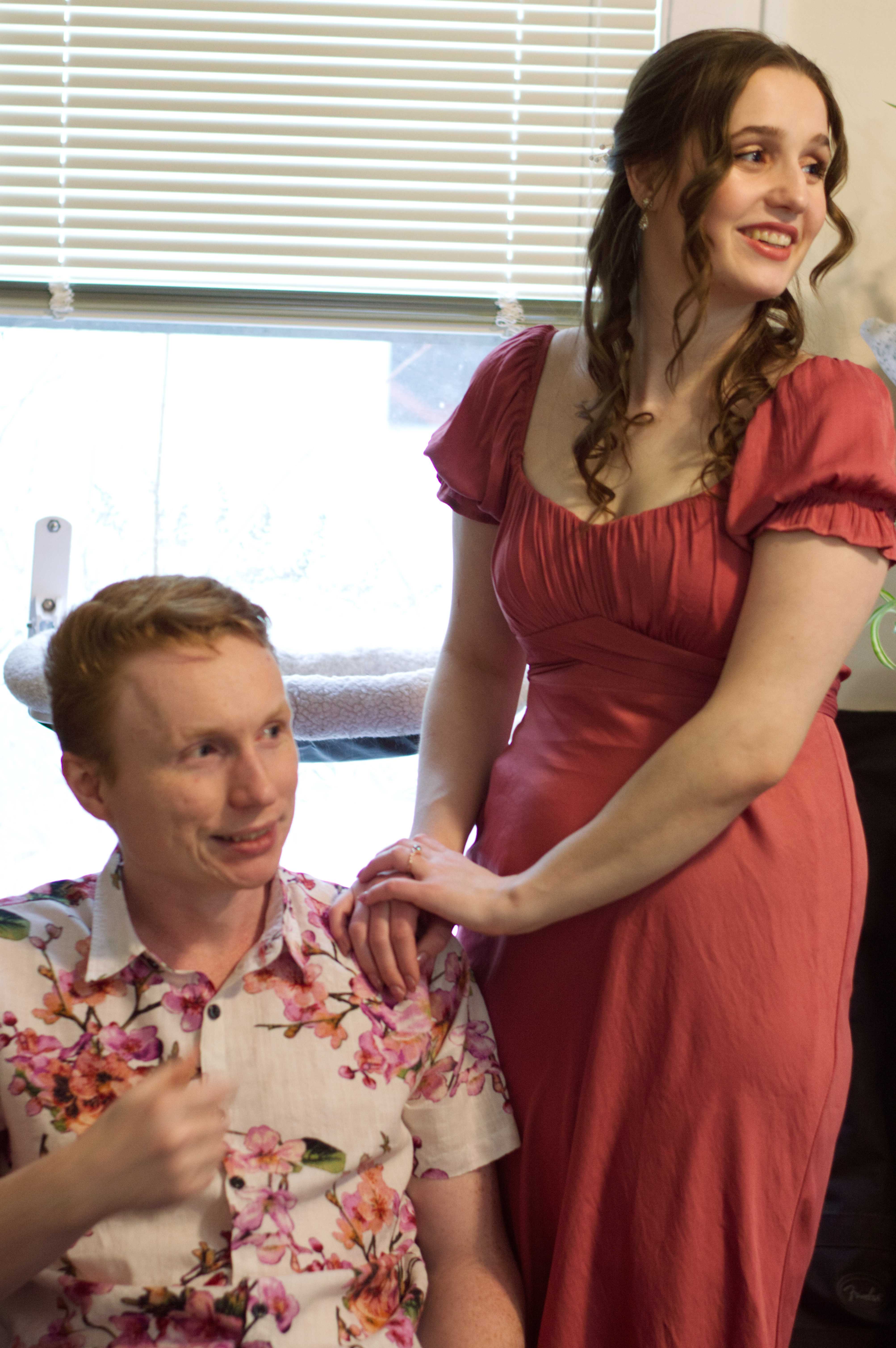 Photo of Mat sitting down with his fiance, Abigail, standing next to him with her hand on his shoulders. Mat is in a white shirt with pink flowers on it, while Abigail is in a pink dress