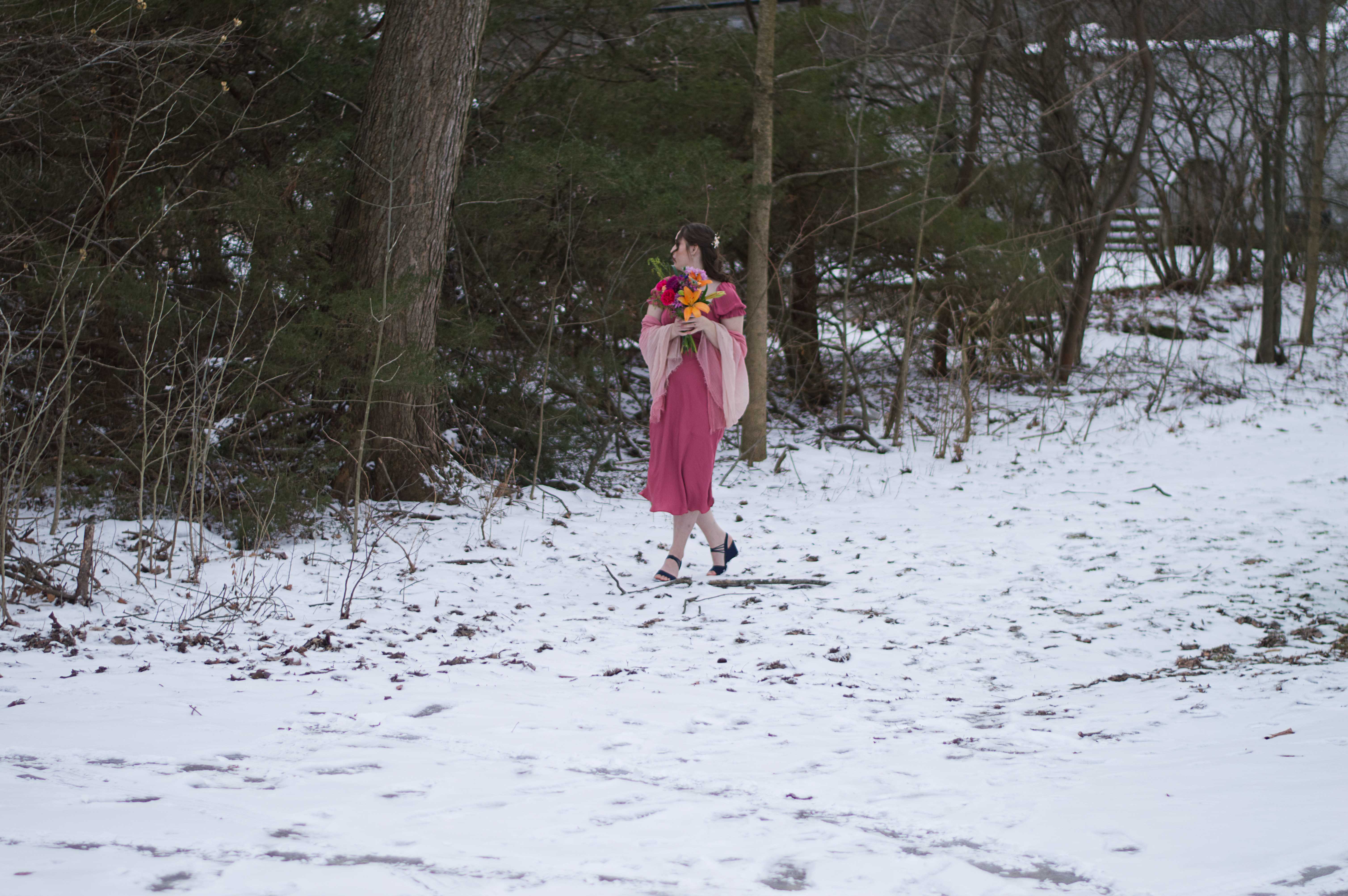 A photo of Abigail in her pink dress, pink shawl, blue heels, and a boquet of flowers she's holding standing in the snow