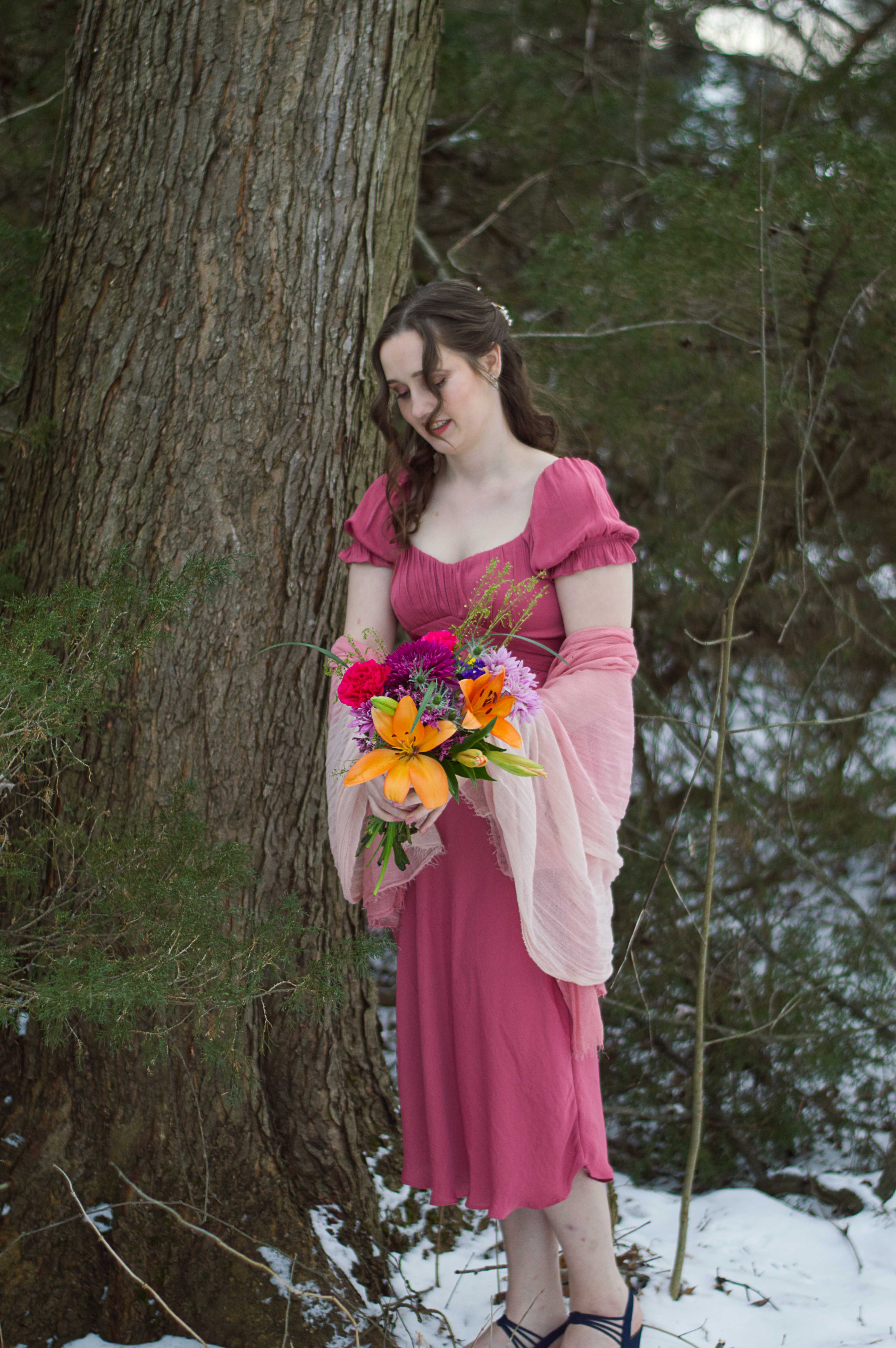 A portrait photo of Abigail standing in her pink dress, pink shawl, blue heels, and a boquet of flowers she's holding standing in the snow