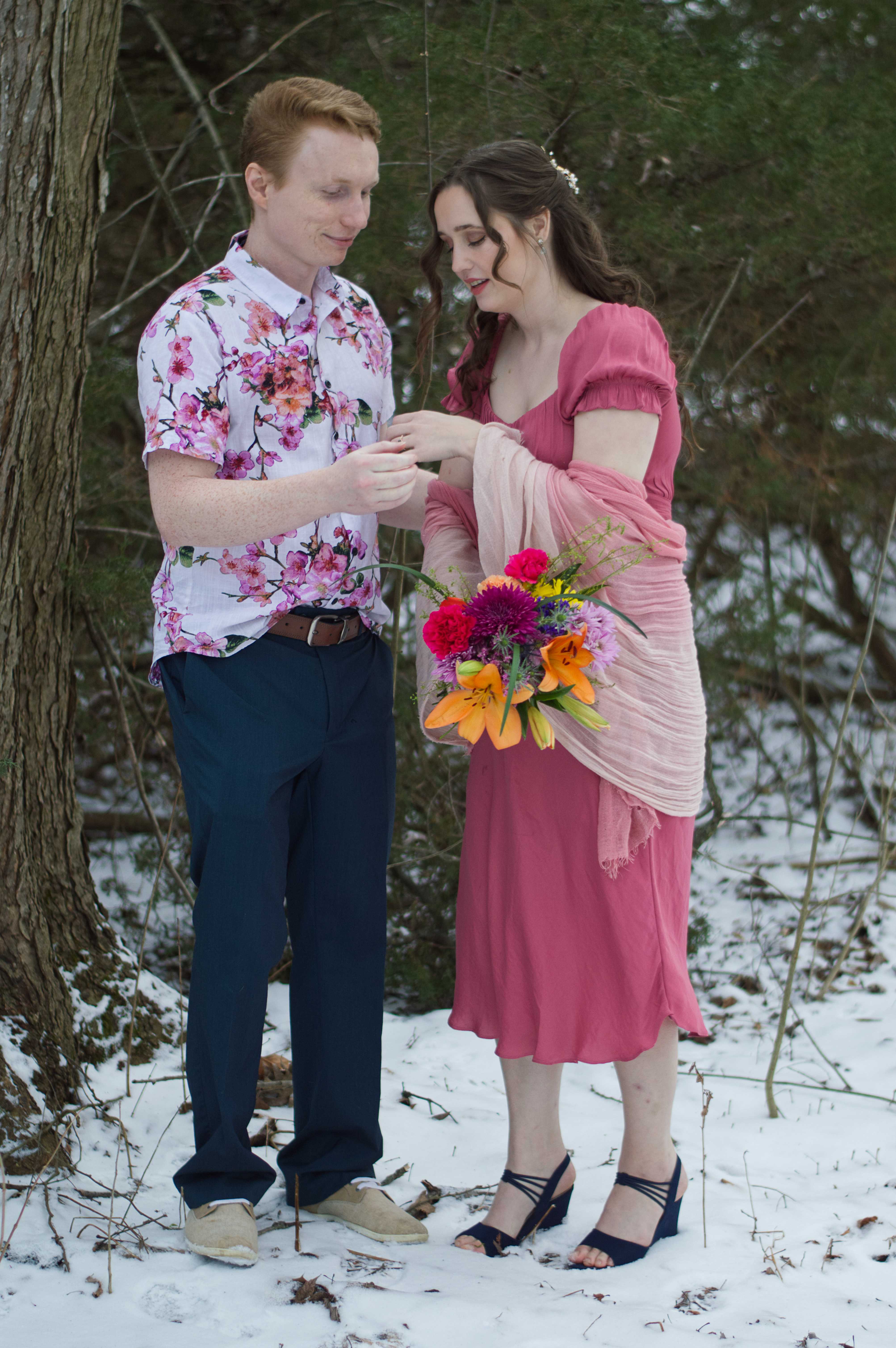 Photo of a man and a woman standing outdoors in the snow. The man (Mat) is wearing a white flowery shirt with blue bottoms, and is helping the woman with a ring on her finger. The woman (Abigail) is in a pink dress, pink shall, heels, and holding a boquet of bright flowers