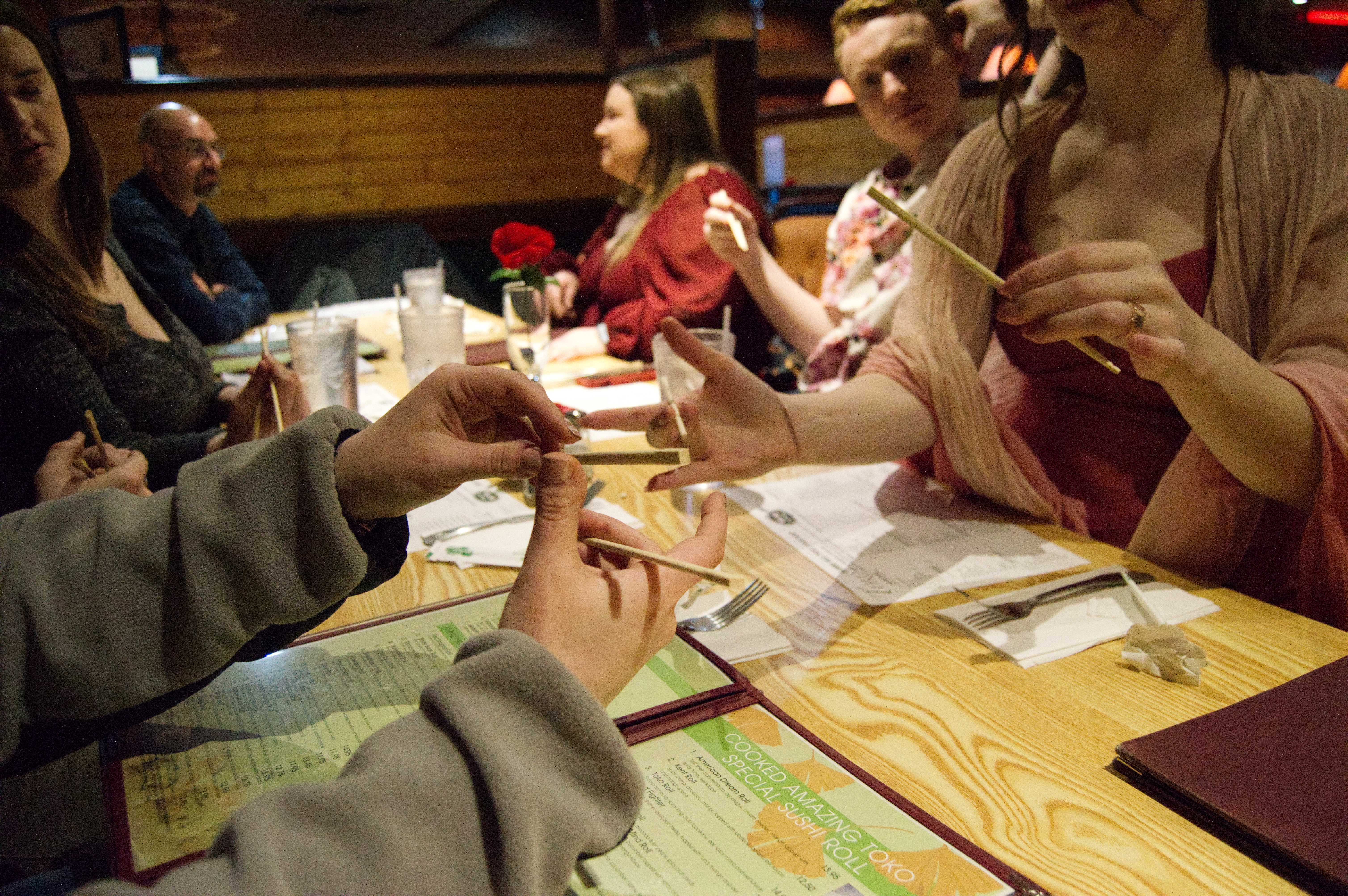 A closeup photo of Michelle trying to hold chopsticks, while Abigail tries to show her what to do, and Mat sits in the background looking on