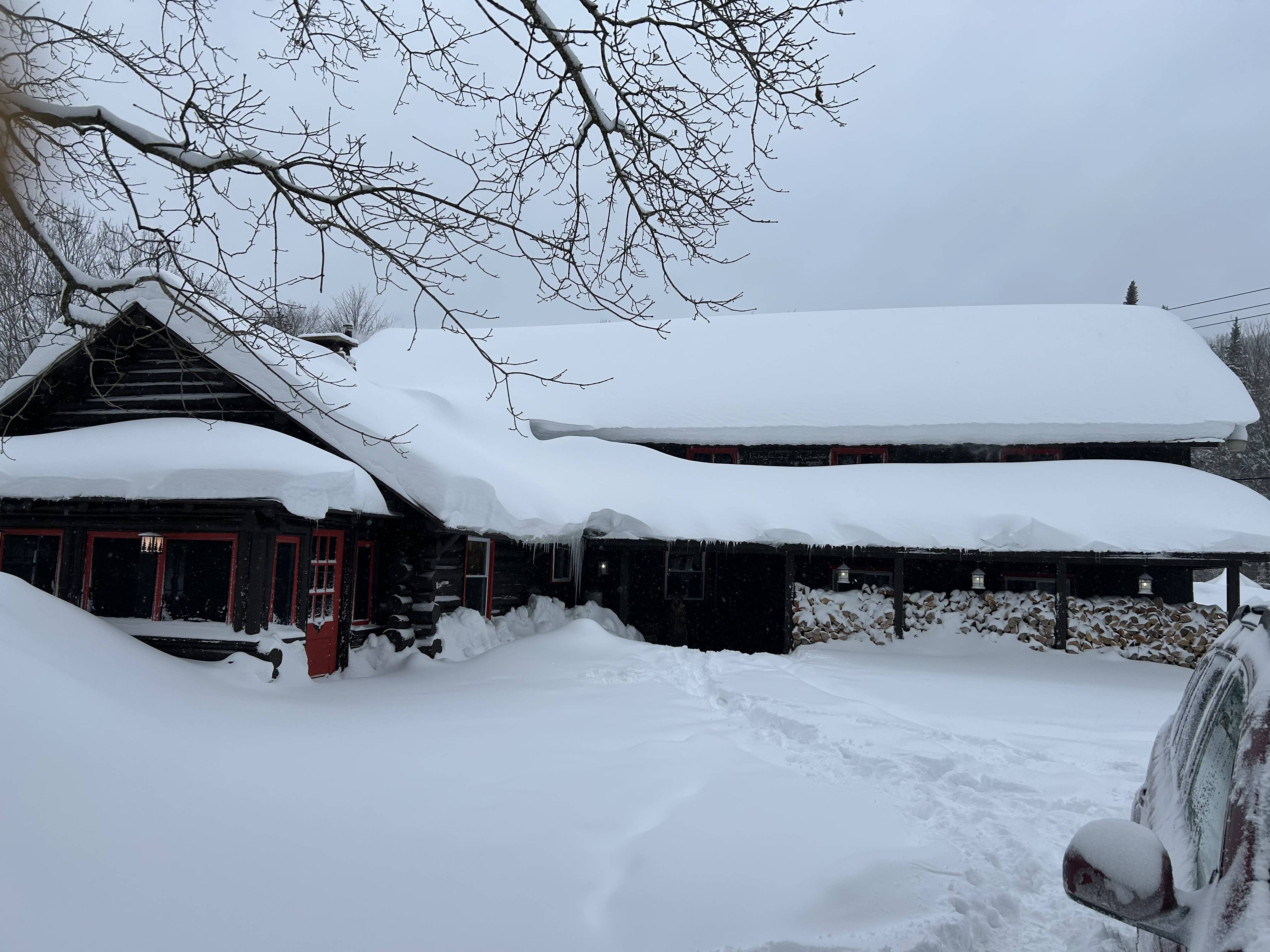 A photo of a snow covered cabin. There is a lot of snow on the ground and roof. The snow was roughly knee deep. You can see some firewood stacked on the outside of the cabin