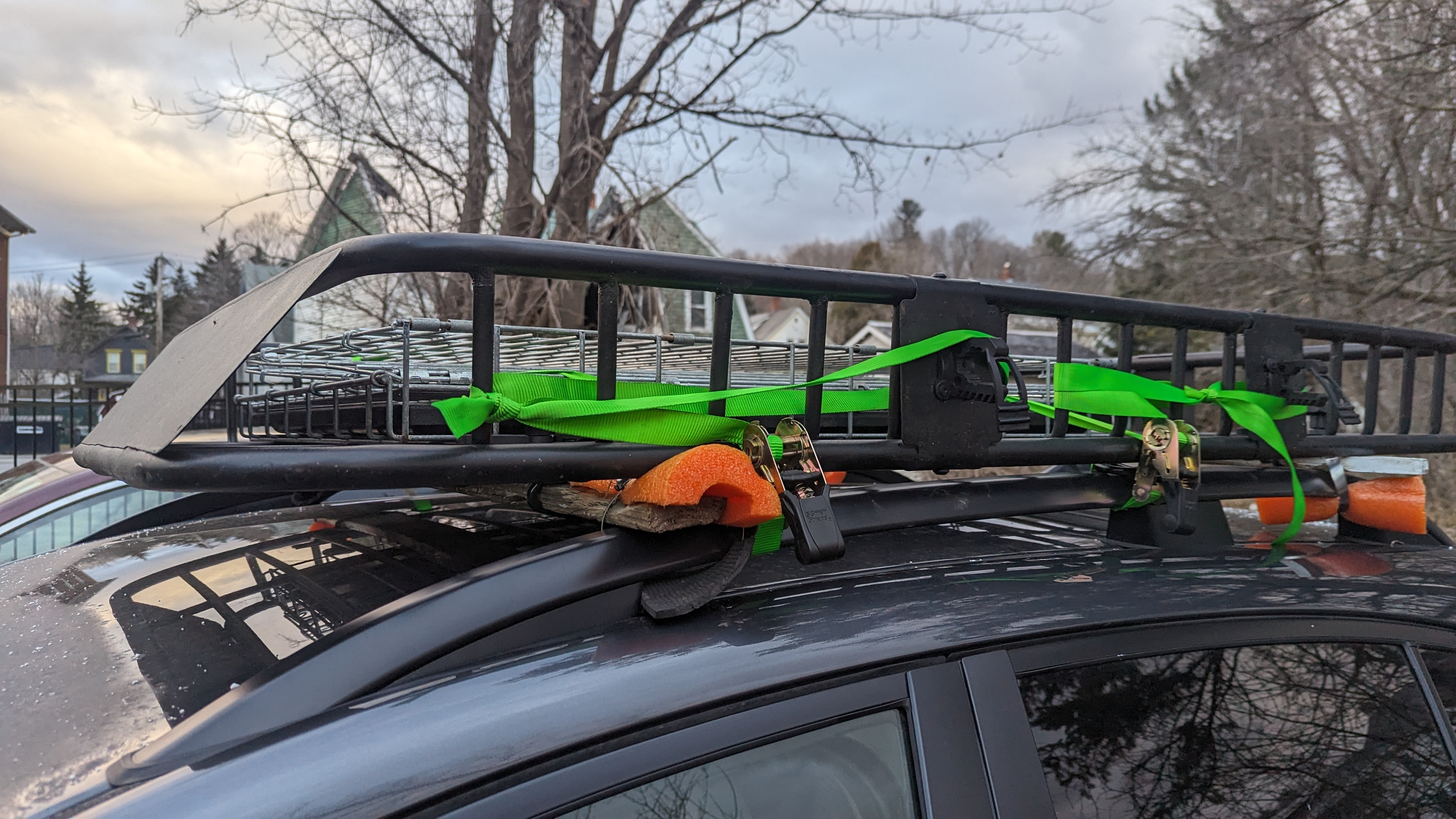 picture of a roof rack on top of a car. Notably the roof rack is being held to the rails with some wooded crossbars, which a basket sits on top of. There are also some green ratchet straps holding everything together