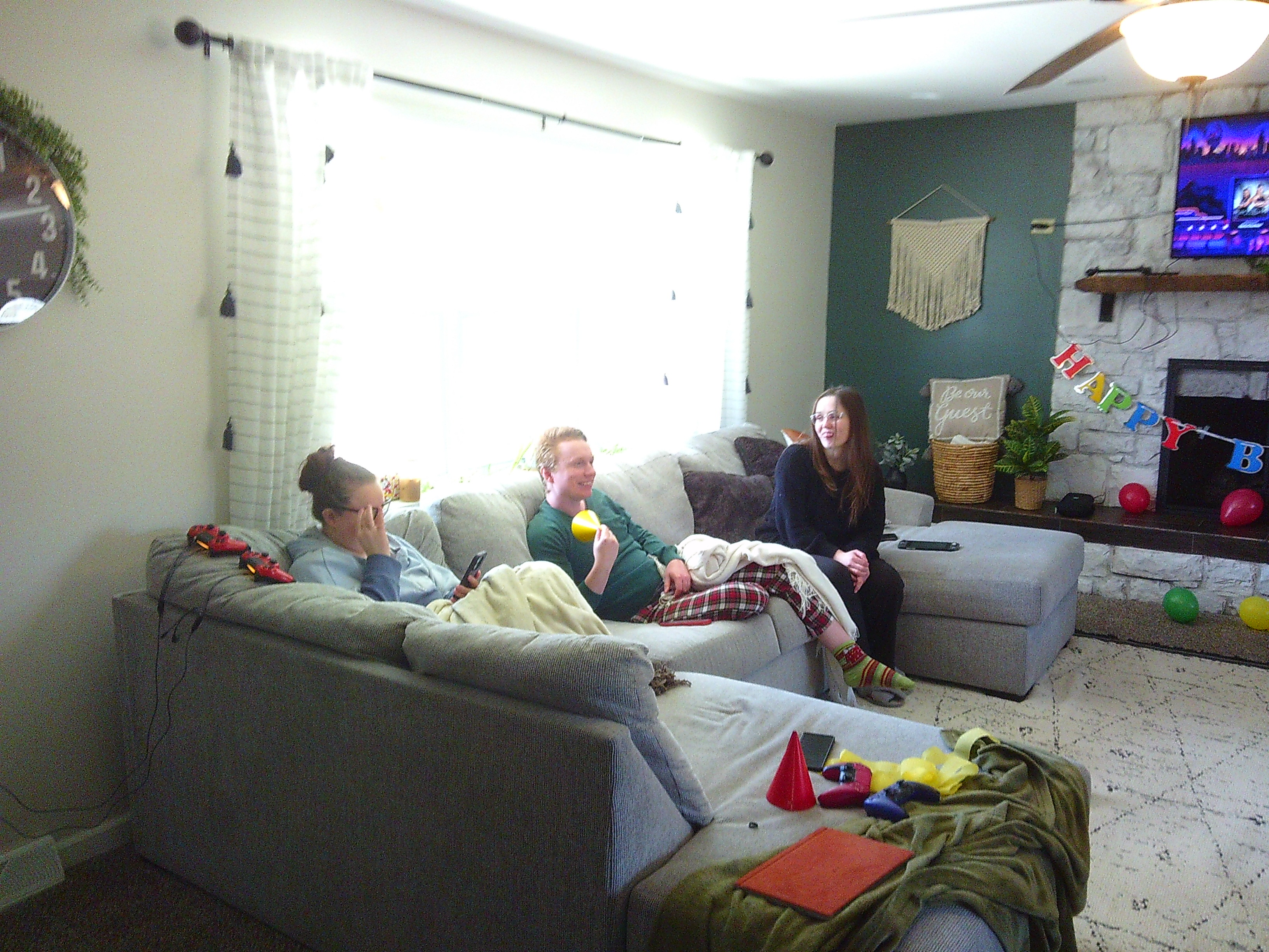 photo of 3 people sitting on a couch. One is on their phone, one is smiling holding a yellow party hat, another is smiling at the one holding the hat