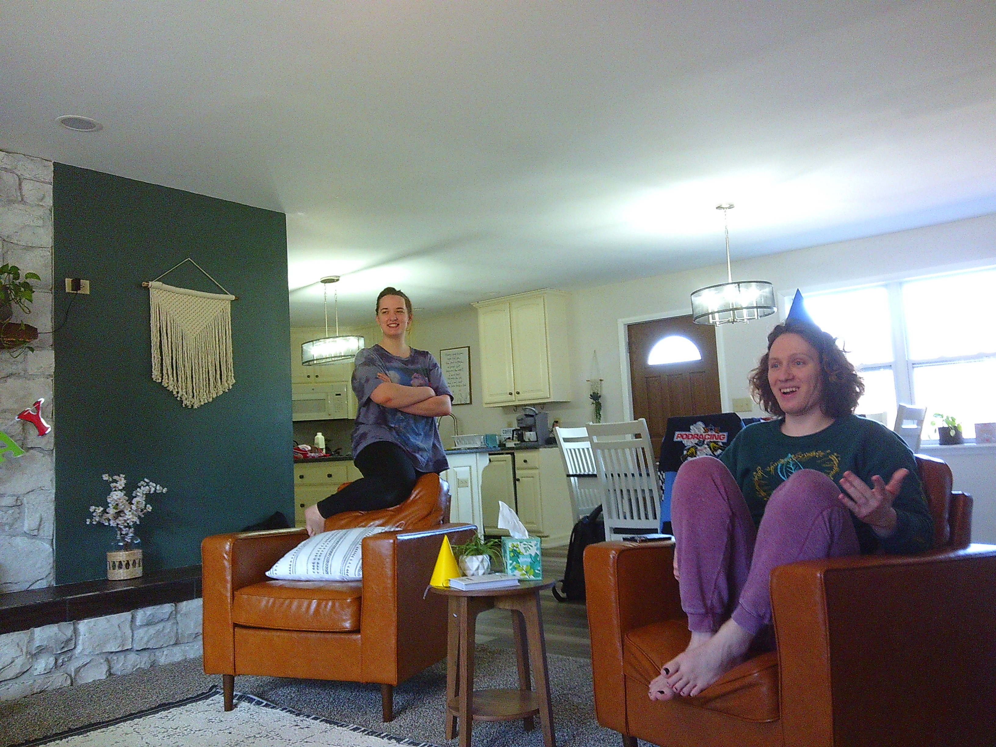 photo of a man in a blue party hat sitting in a chair, talking to someone off camera. There is a woman with her leg around a chair, arms crossed, also looking off camera at someone