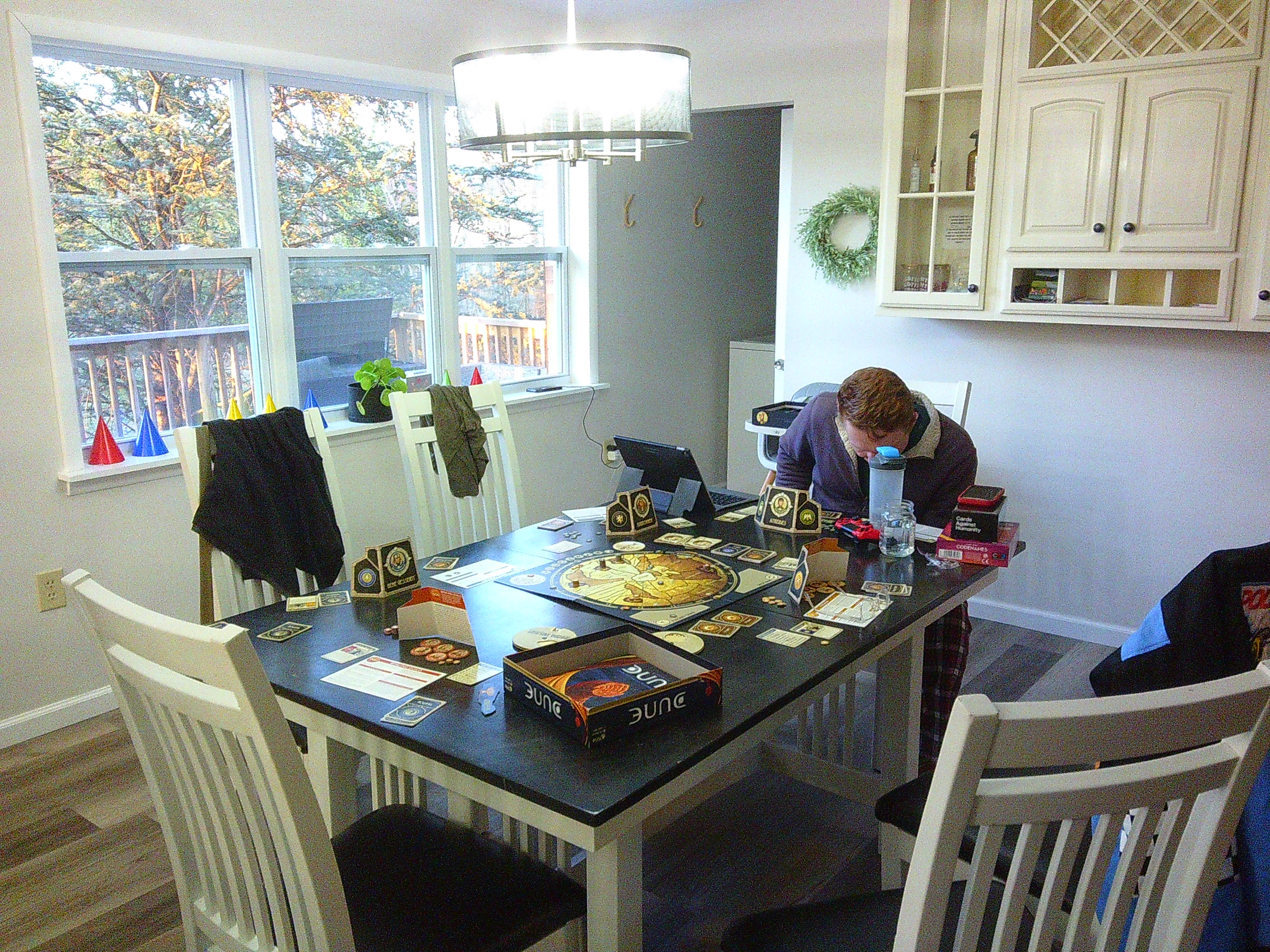 photo of a man at a kitchen table looking down at his phone. On the table the board game Dune has been setup, and there are pieces everywhere