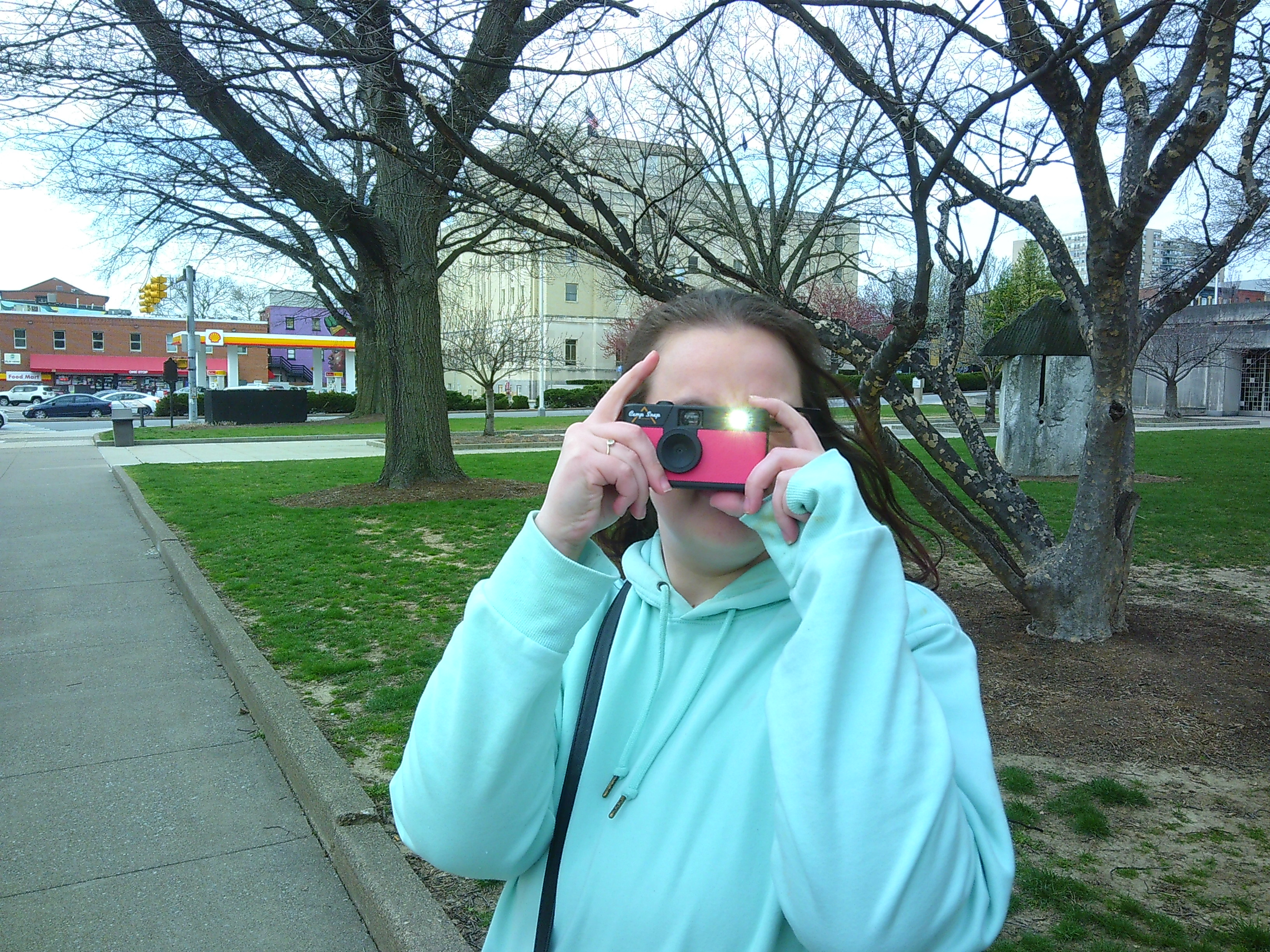 Picture of a woman holding a pink camera taking a photo of the person taking a photo of her. She is wearing a blue sweatshirt