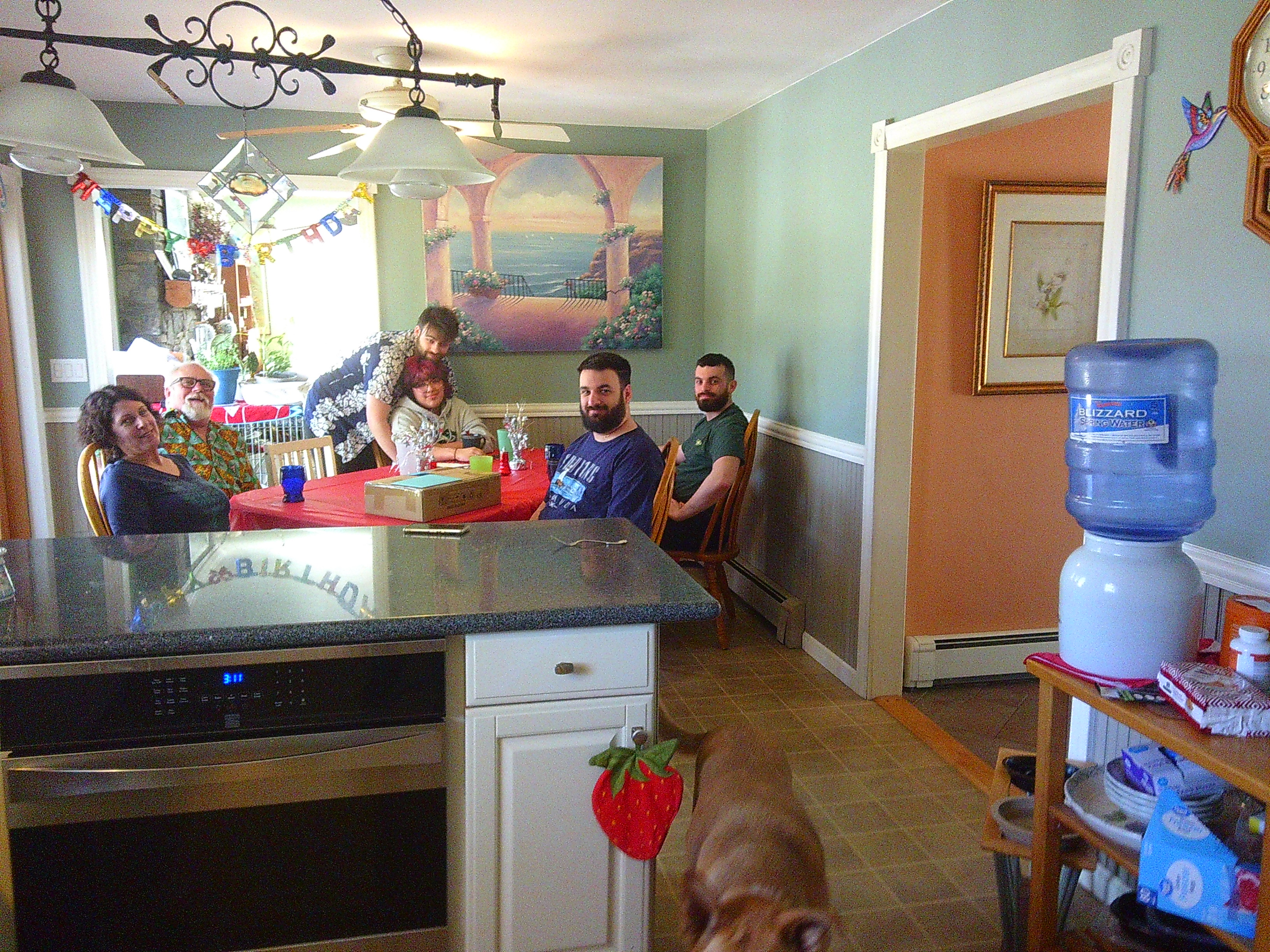 Picture of a bunch of people around a table in a kitchen. You can see a Happy Birthday banner in the distance. There is a dog half out of the frame towards the bottom of the photo.